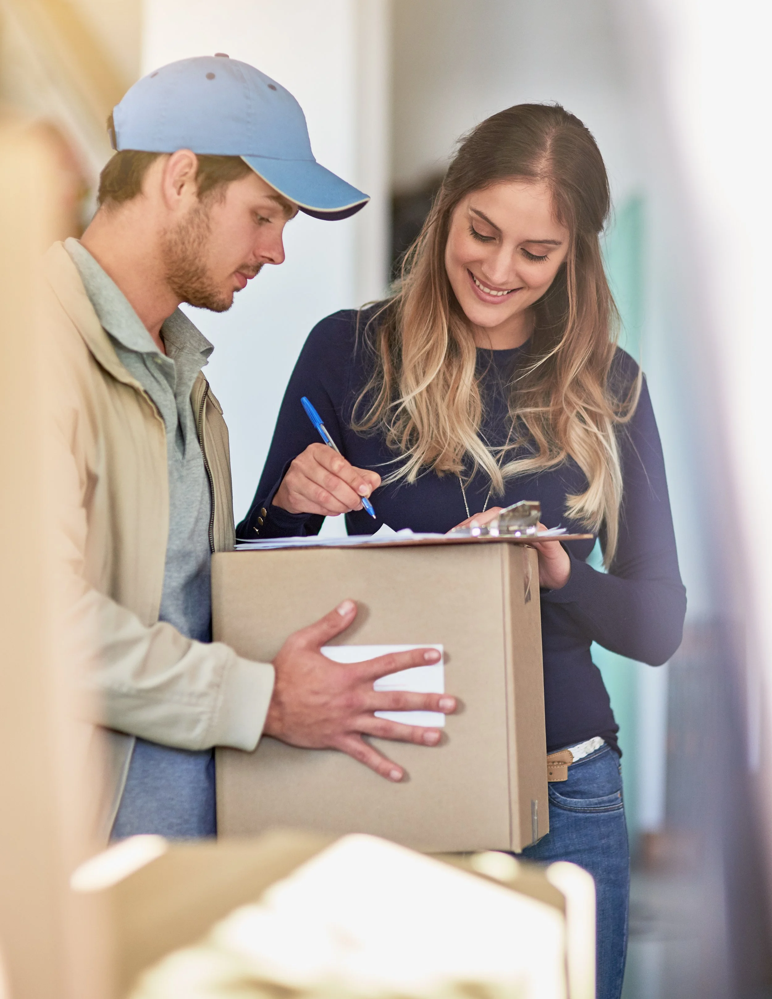 A young man and woman look at a clipboard and a box, with the man holding the box and the woman writing on the clipboard, in an indoor setting.