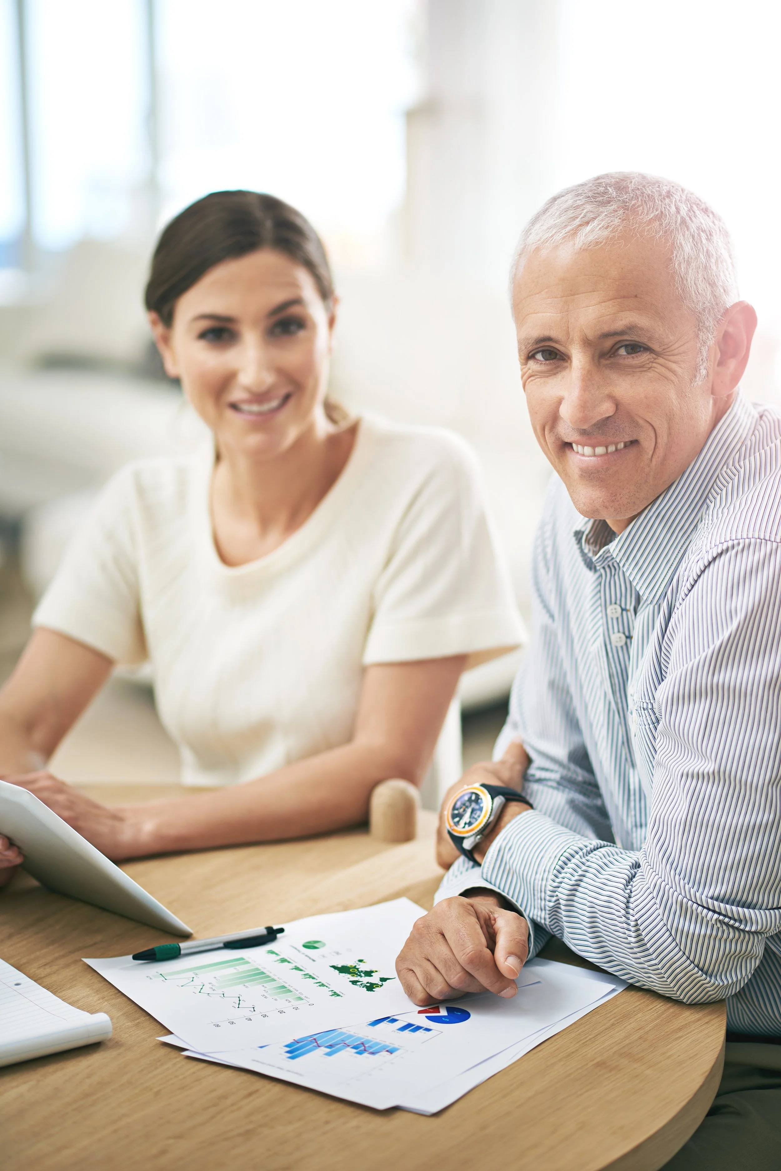 Two professionals, a woman and a man, smiling at a meeting table with charts and a tablet.