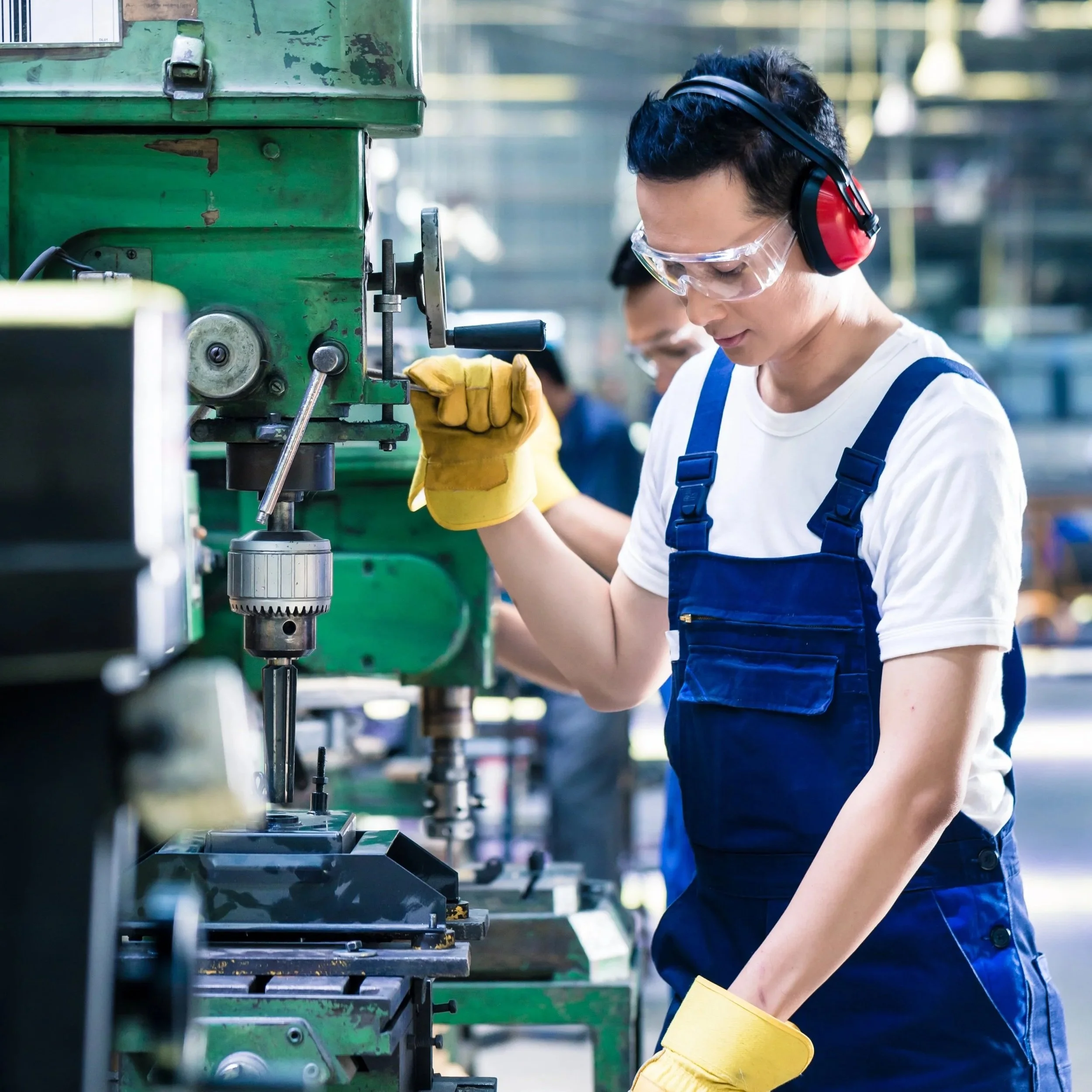 Worker wearing safety glasses and earmuffs, operating a green industrial milling machine in a factory setting.