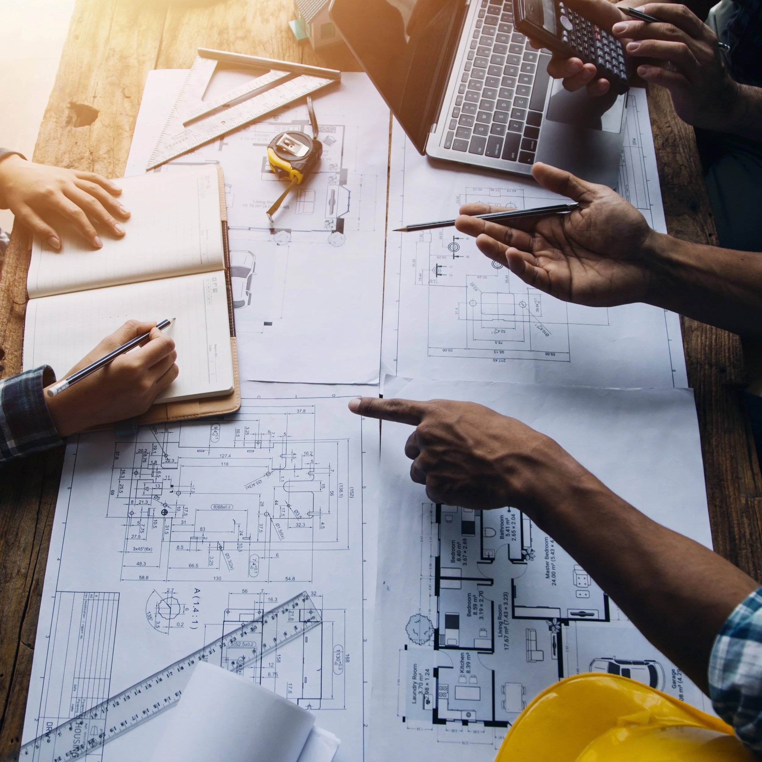People working on architectural blueprints and plans at a table with a laptop, ruler, tape measure, notebook, and calculator.