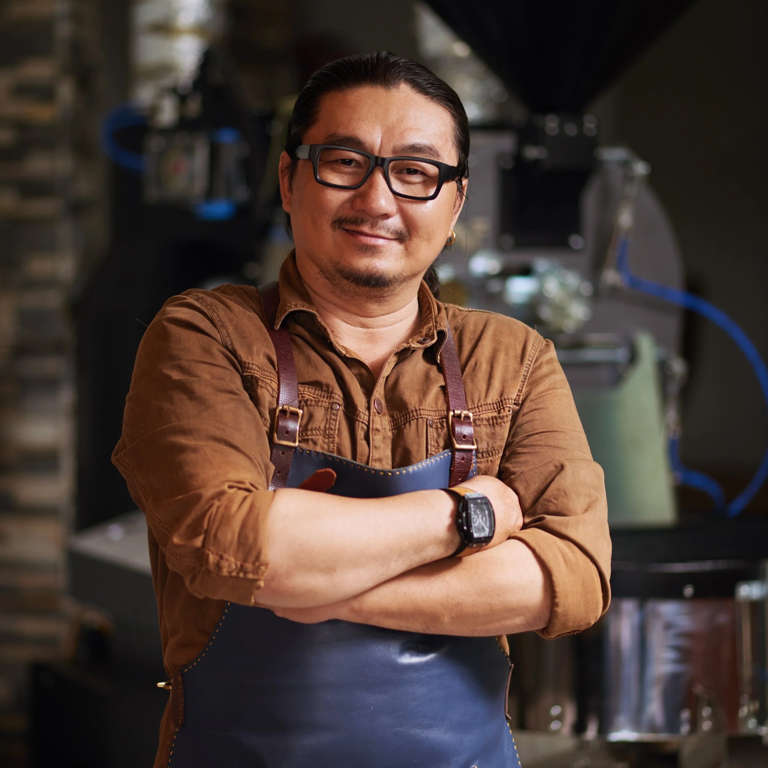 A man with glasses and facial hair, wearing a brown shirt and a leather apron, stands with arms crossed in a kitchen or workshop environment.