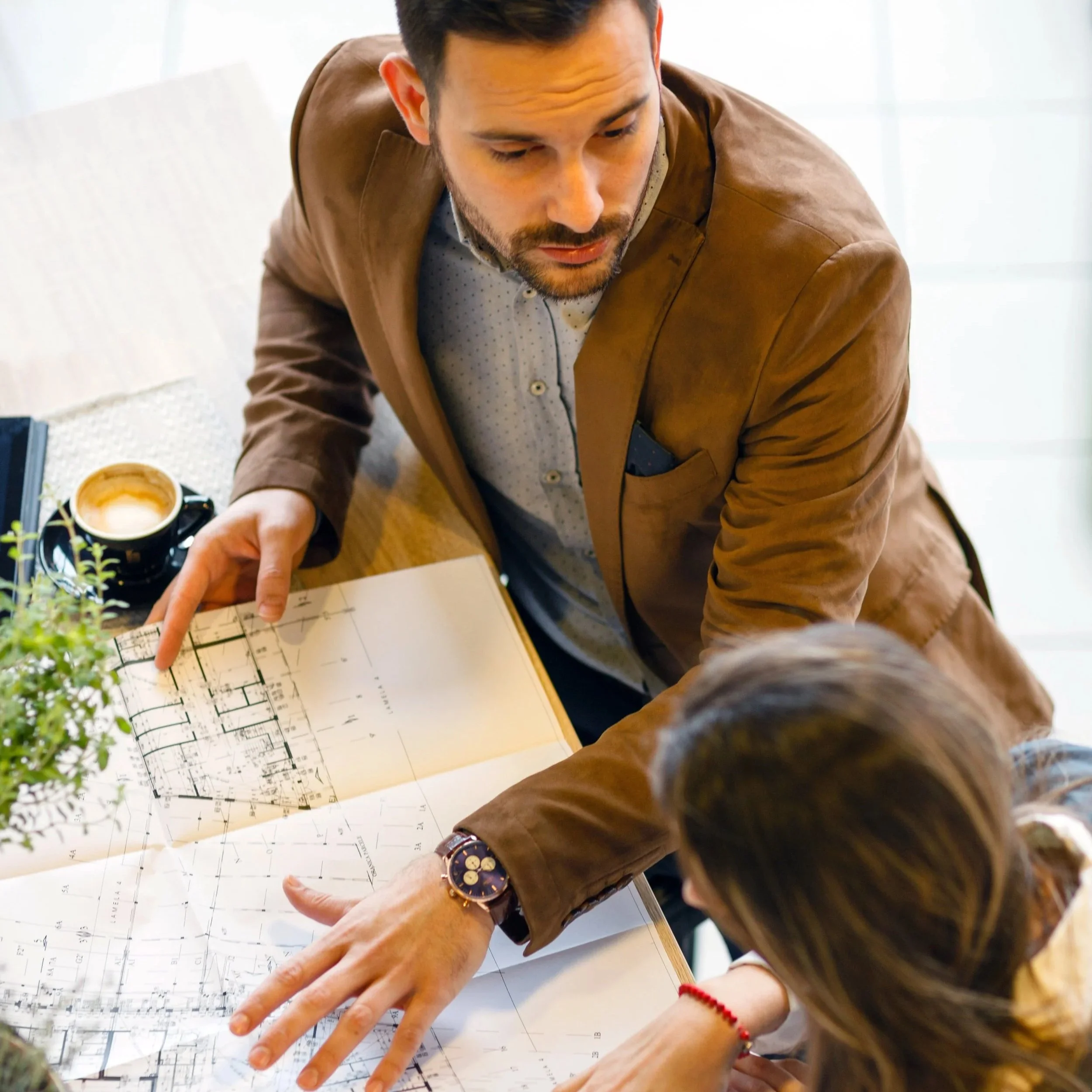 A man in a brown jacket and a patterned shirt points to architectural plans on a table, while a woman with a red bracelet listens. There is a cup of coffee, a plant, and a black object nearby.
