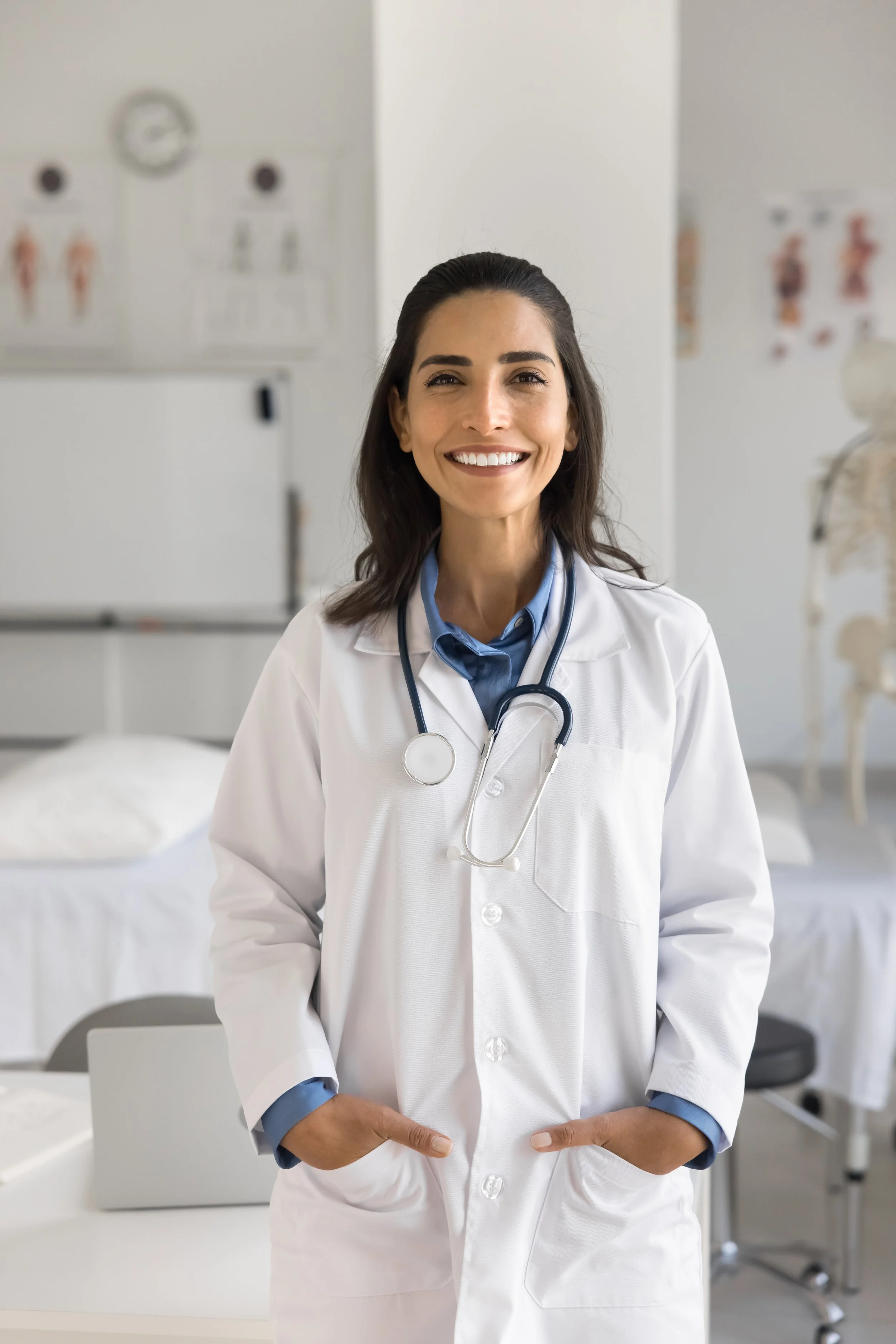 A smiling female doctor with a stethoscope around her neck, standing in a medical office or clinic, with medical charts and anatomical models in the background.