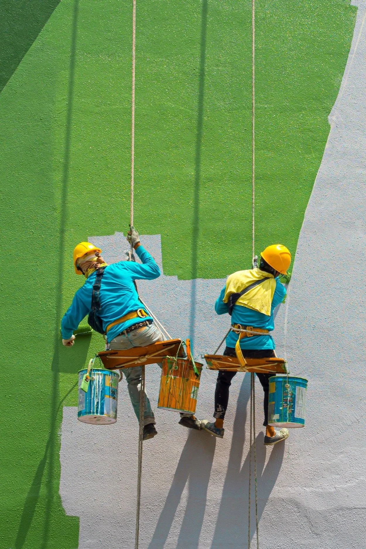 Two workers wearing yellow safety helmets and harnesses are painting a wall green. They are seated on wooden platforms hanging by ropes, with paint buckets attached to their belts.
