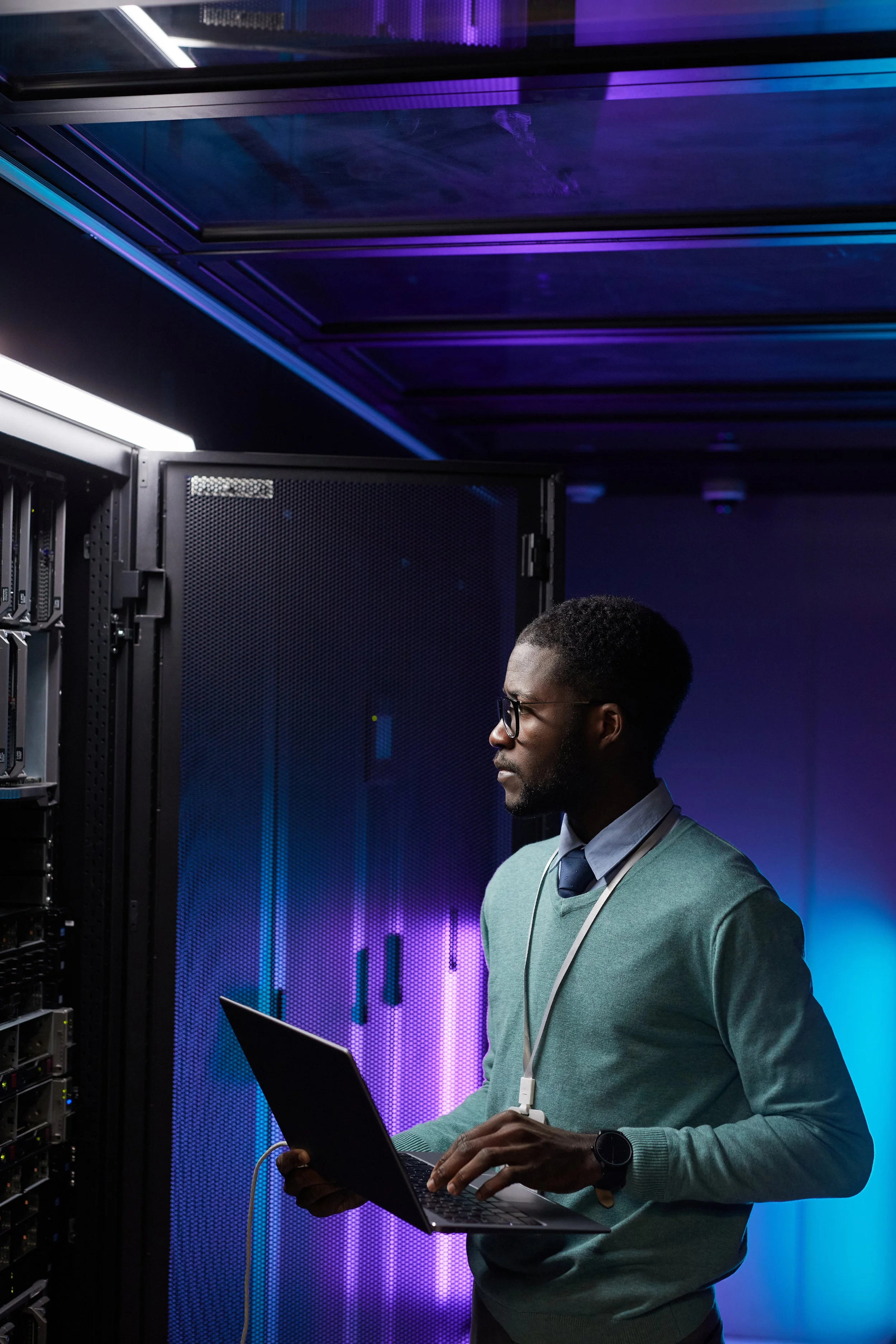 A man with glasses and a teal sweater holding a laptop standing in front of a server rack in a data center with purple and blue lighting.