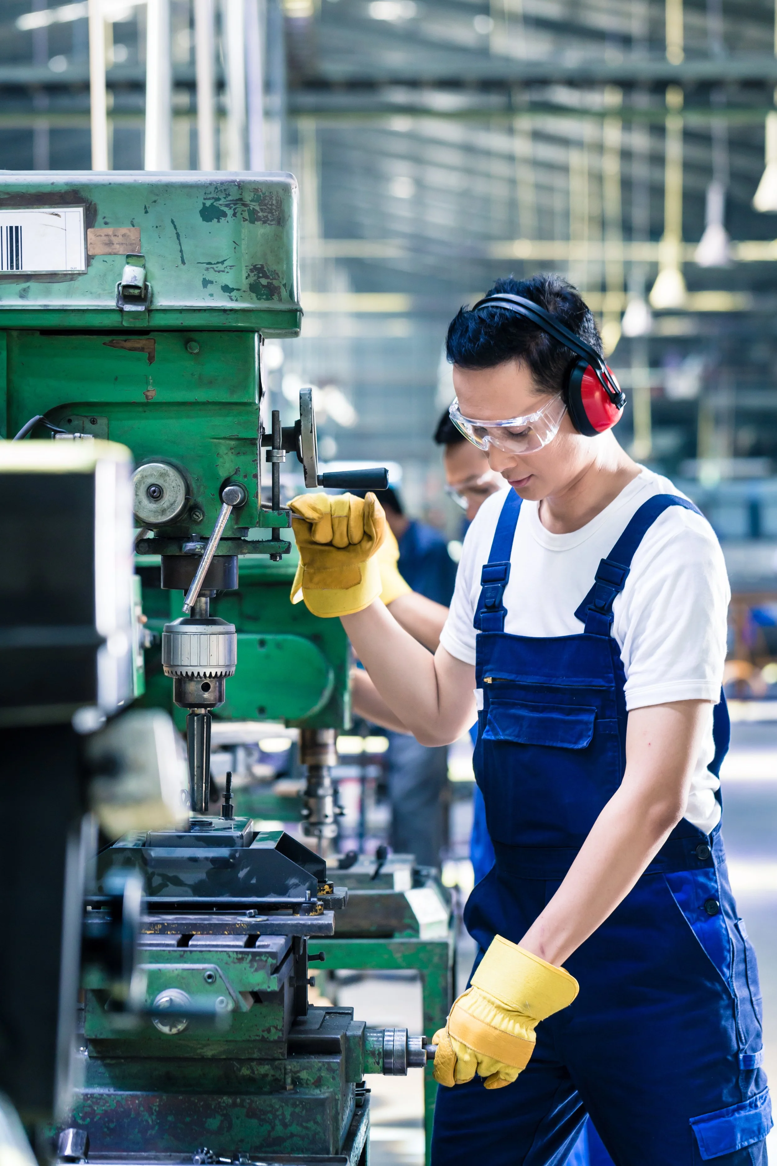A woman wearing safety goggles, ear protection, yellow gloves, a white t-shirt, and blue overalls is operating a green industrial machine in a factory or workshop.