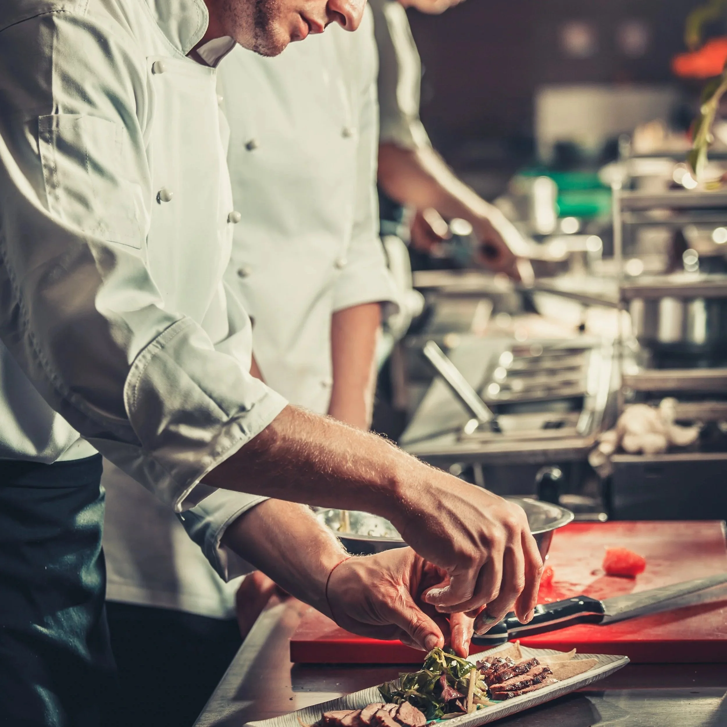 Chef plating food in a professional kitchen with other chefs in the background.