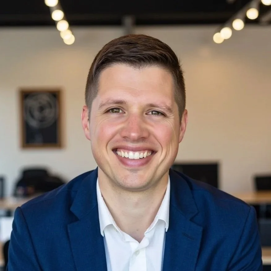 Portrait of a smiling man wearing a dark suit, white shirt, and dark blue tie, with a dark background.