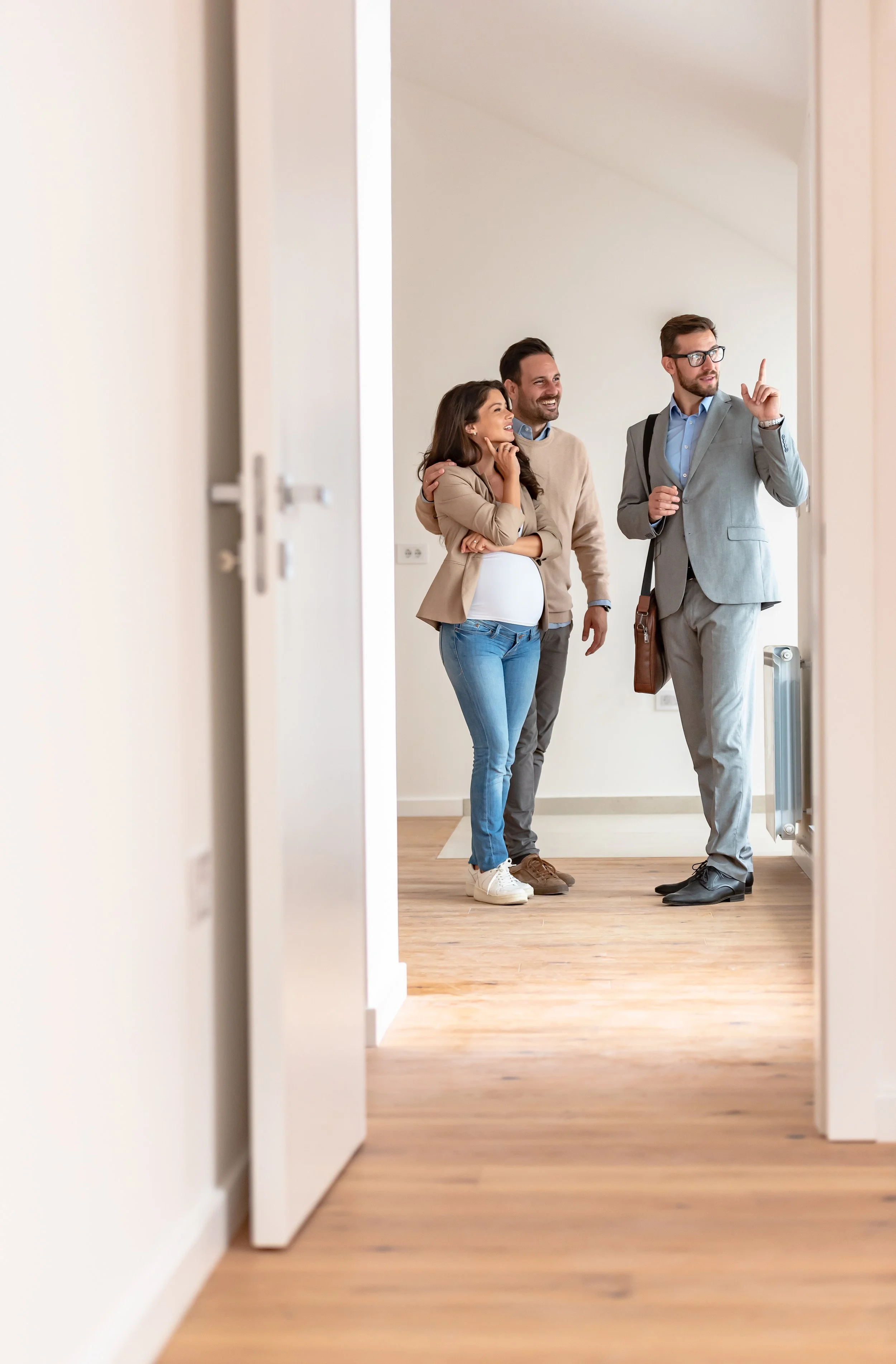 Three people, two men and one woman, standing in a doorway inside a home, engaging in conversation. The woman appears pregnant, and one man is wearing glasses and a grey suit, with a backpack.