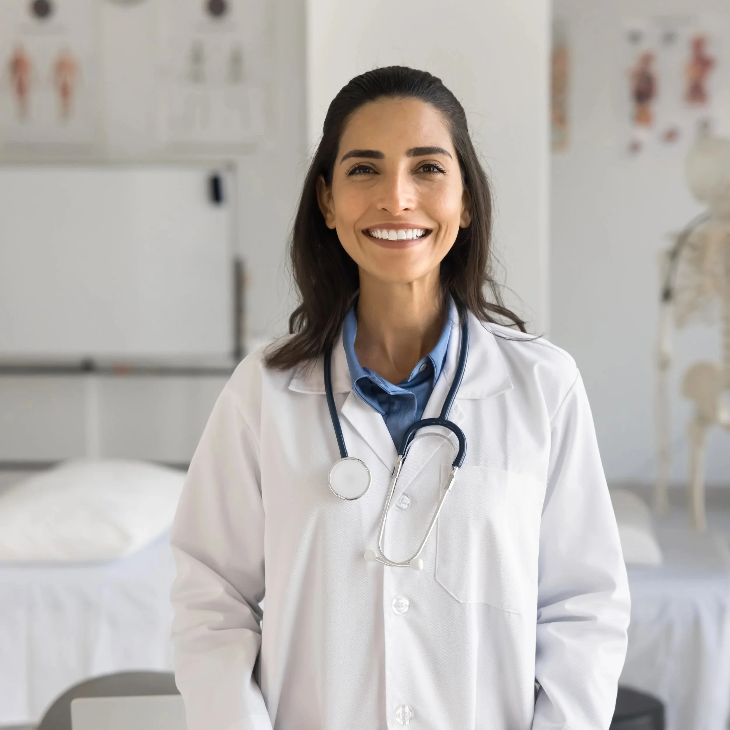 A smiling female doctor in a white coat with a stethoscope around her neck, standing in a medical examination room with medical posters and a skeleton model in the background.