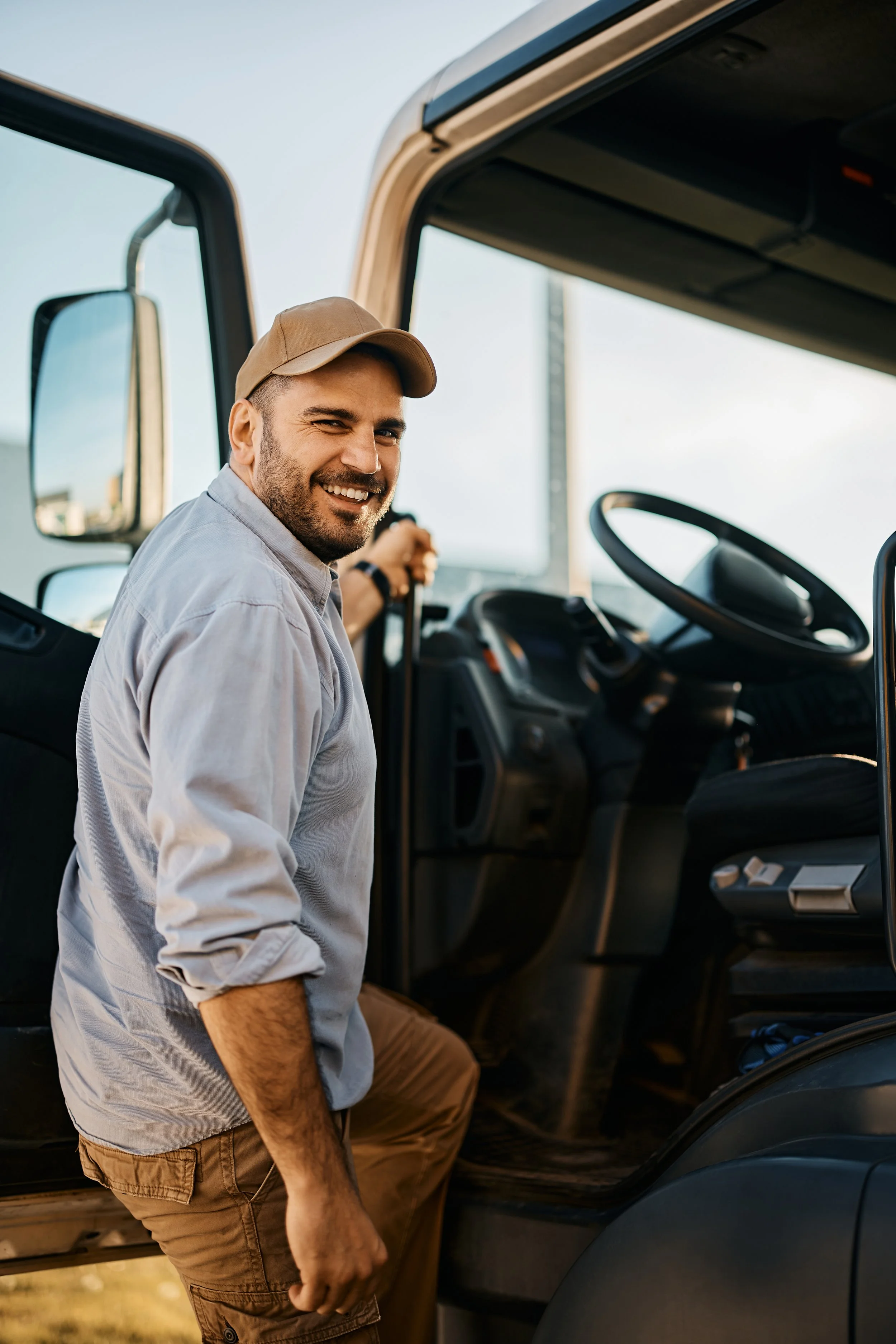 A man smiling and looking at camera while standing by the open door of a semi-truck during daytime.
