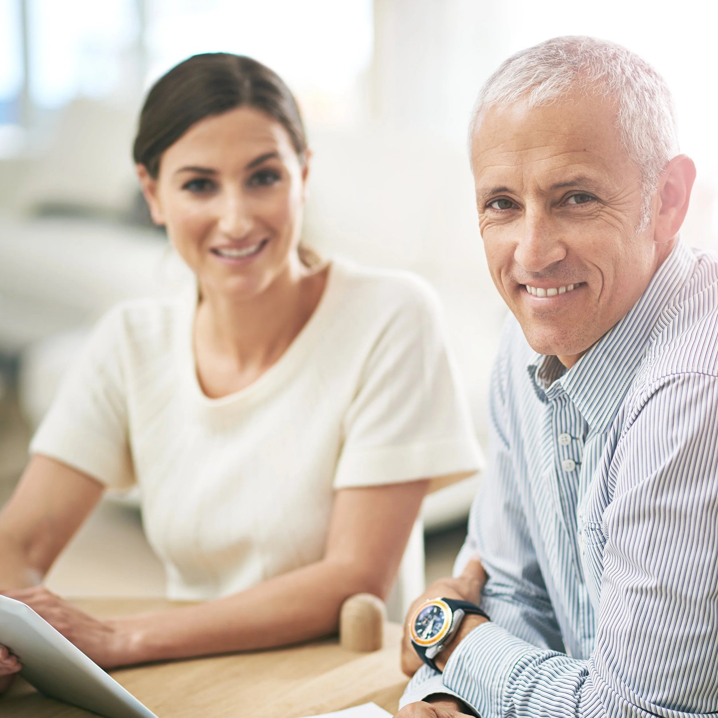 A smiling middle-aged man with short gray hair and a woman with dark brown hair sitting at a table, smiling, with a blurred bright background.