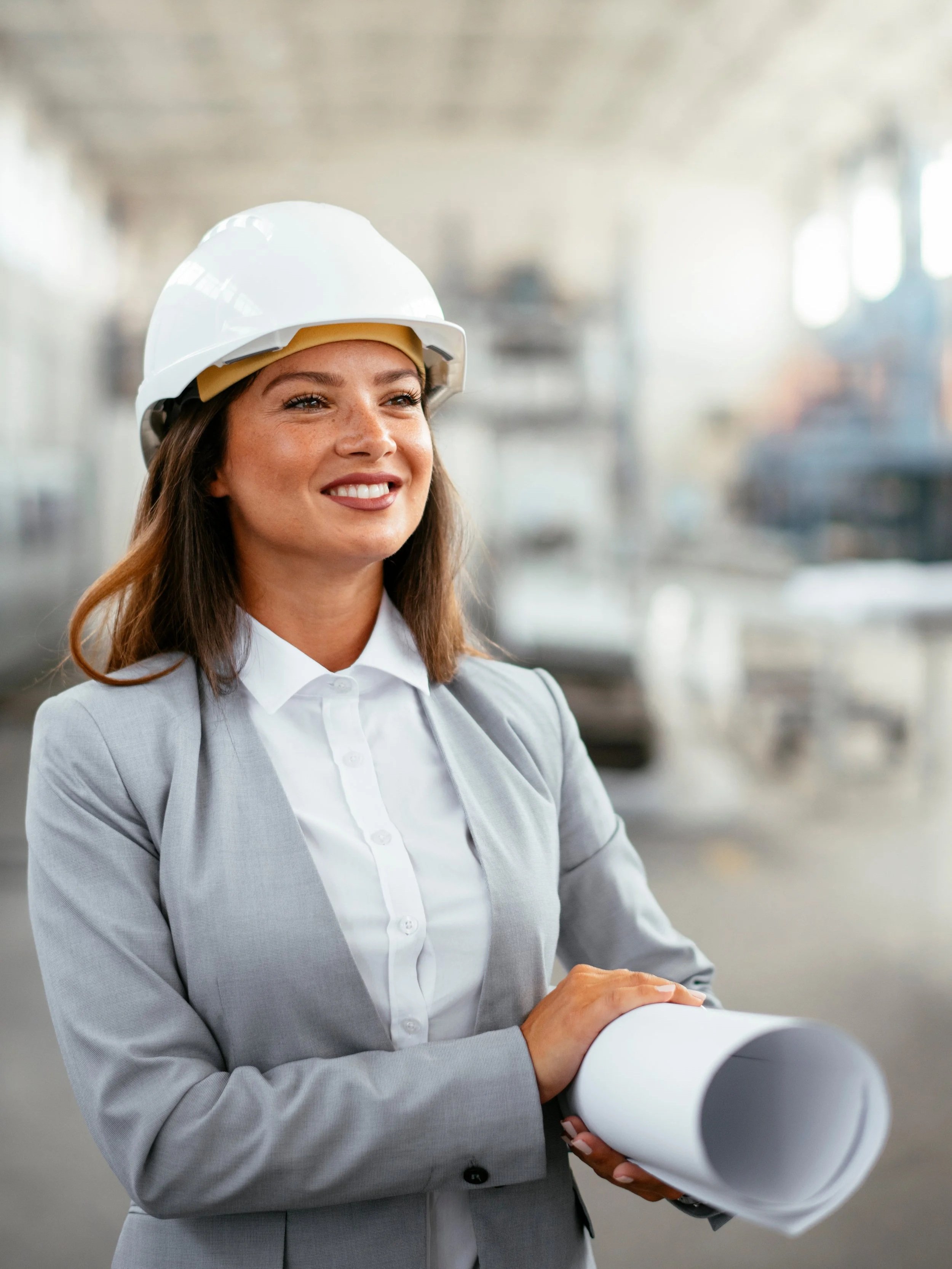 A woman wearing a white safety helmet and business attire, holding a rolled-up blueprint in an industrial or construction setting, smiling confidently.