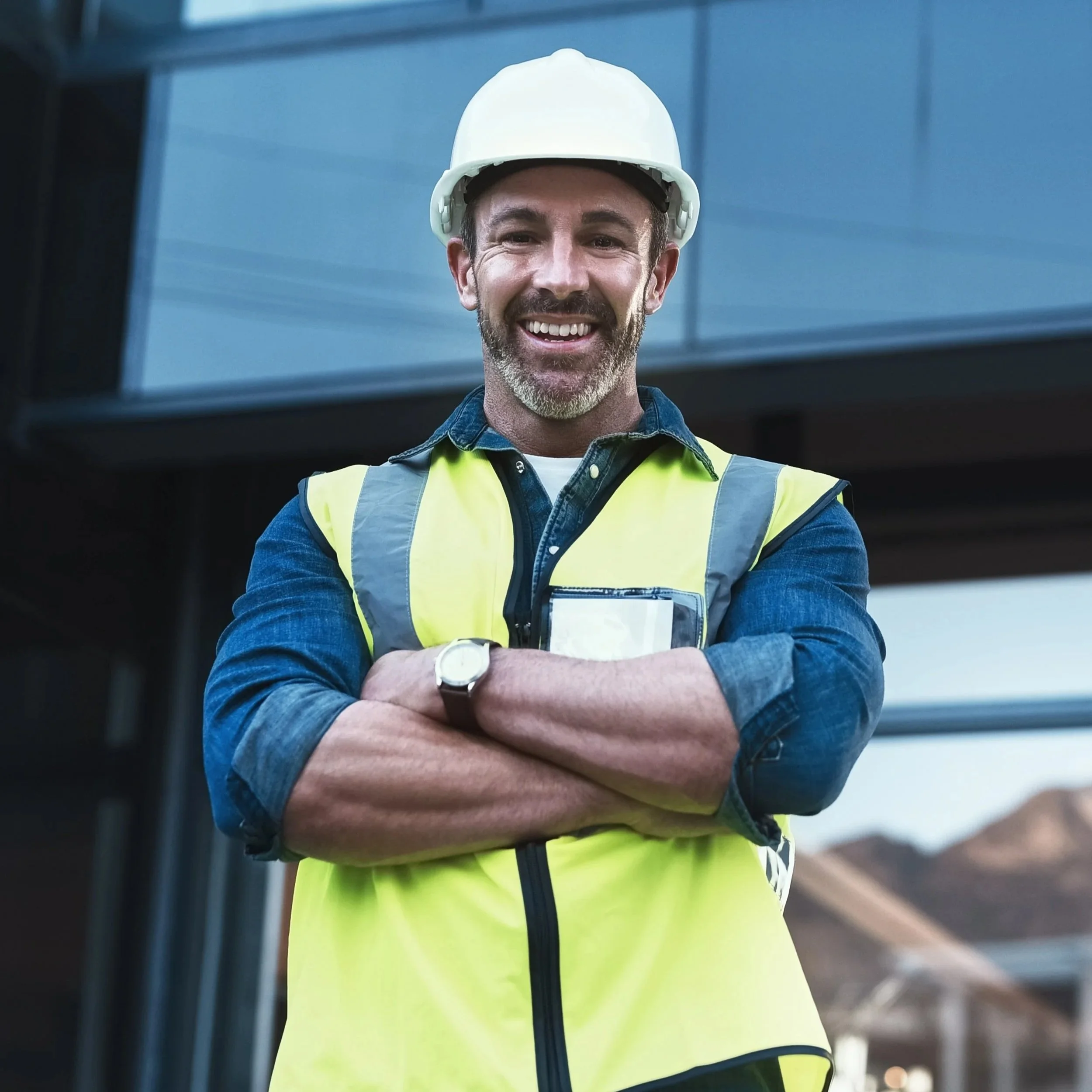 A smiling construction worker wearing a white hard hat, yellow safety vest, and a blue denim shirt with rolled-up sleeves, standing outdoors with arms crossed.