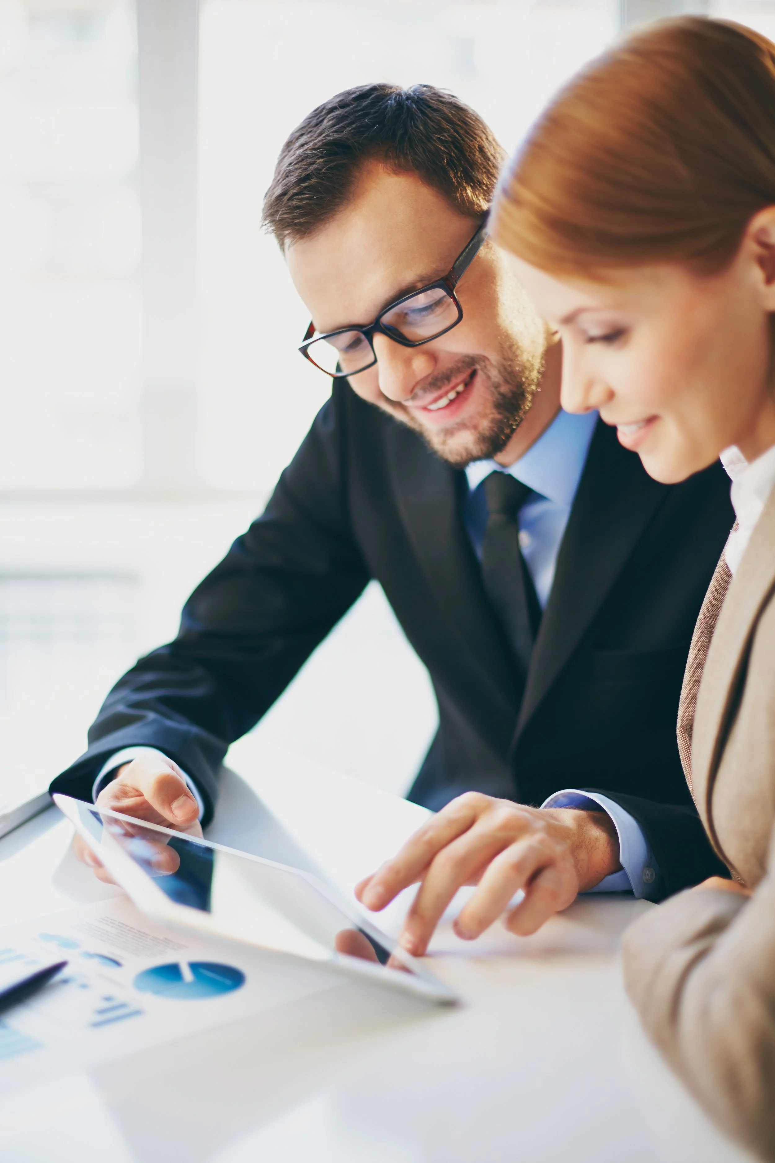 Two business professionals, a man and a woman, sitting at a white table reviewing charts and data on a tablet in a bright office.