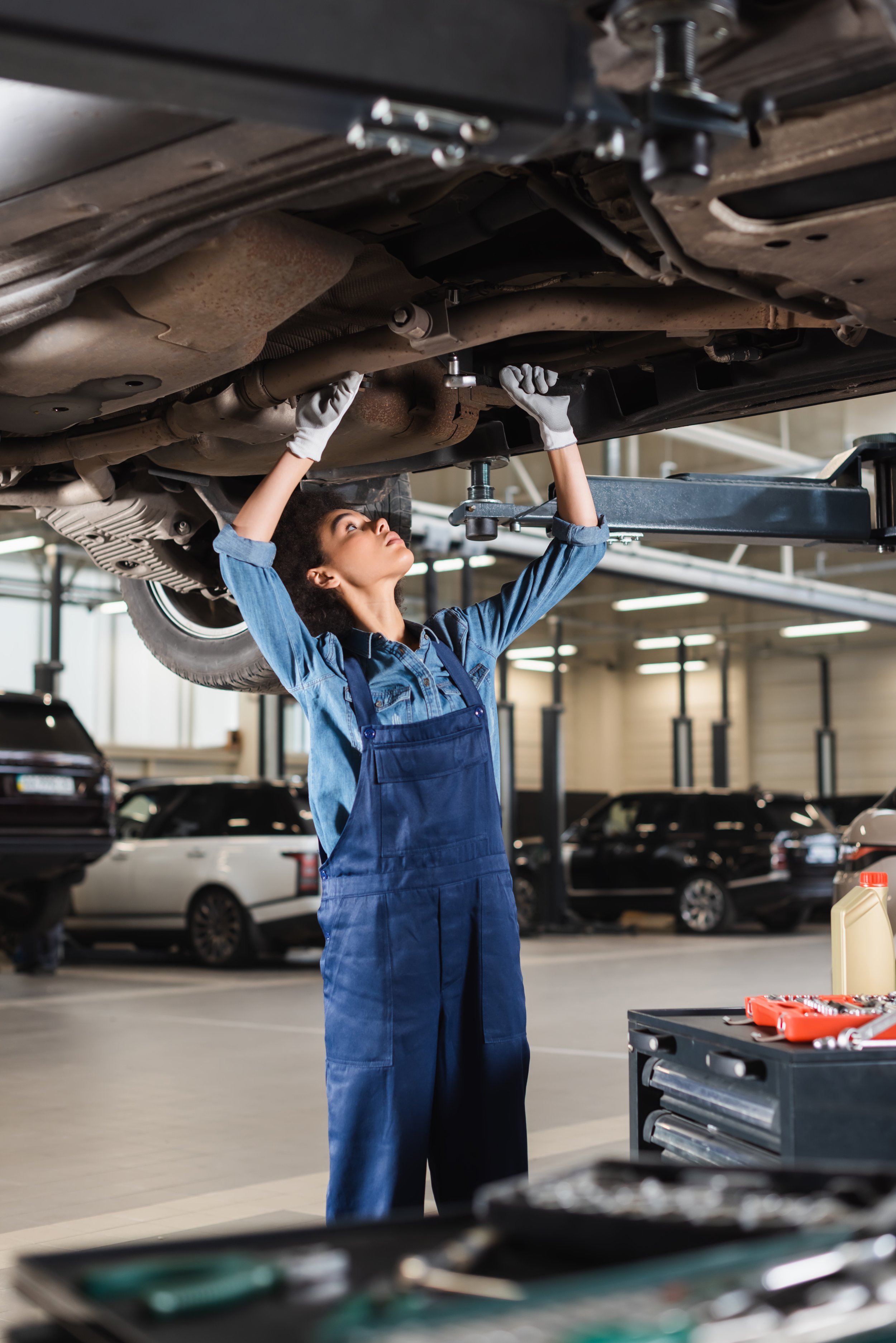 A woman working underneath a car lifted on a hydraulic lift in an auto repair shop, wearing blue coveralls and gloves, with various tools nearby.