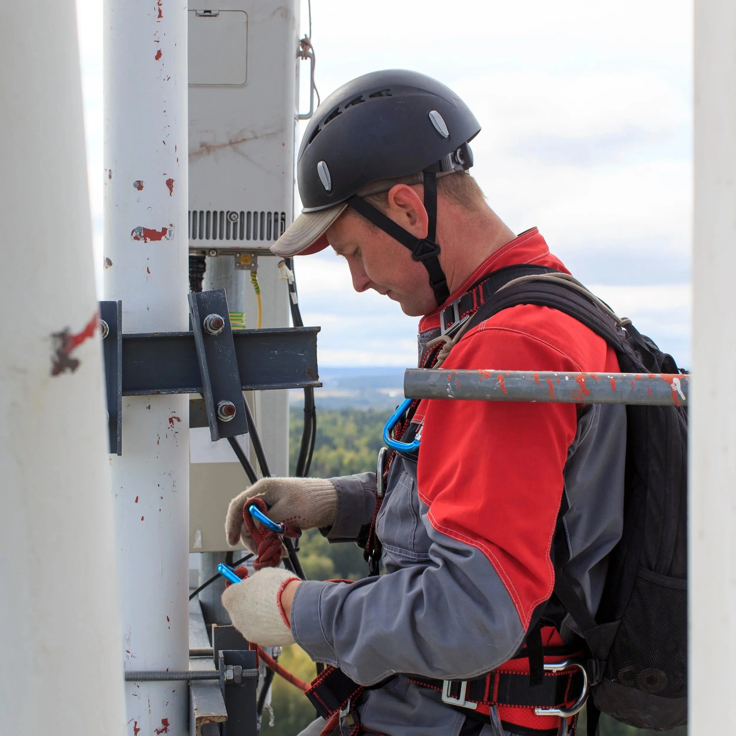 A man in safety gear, including a helmet and gloves, working on electrical components outdoors on a metal structure.