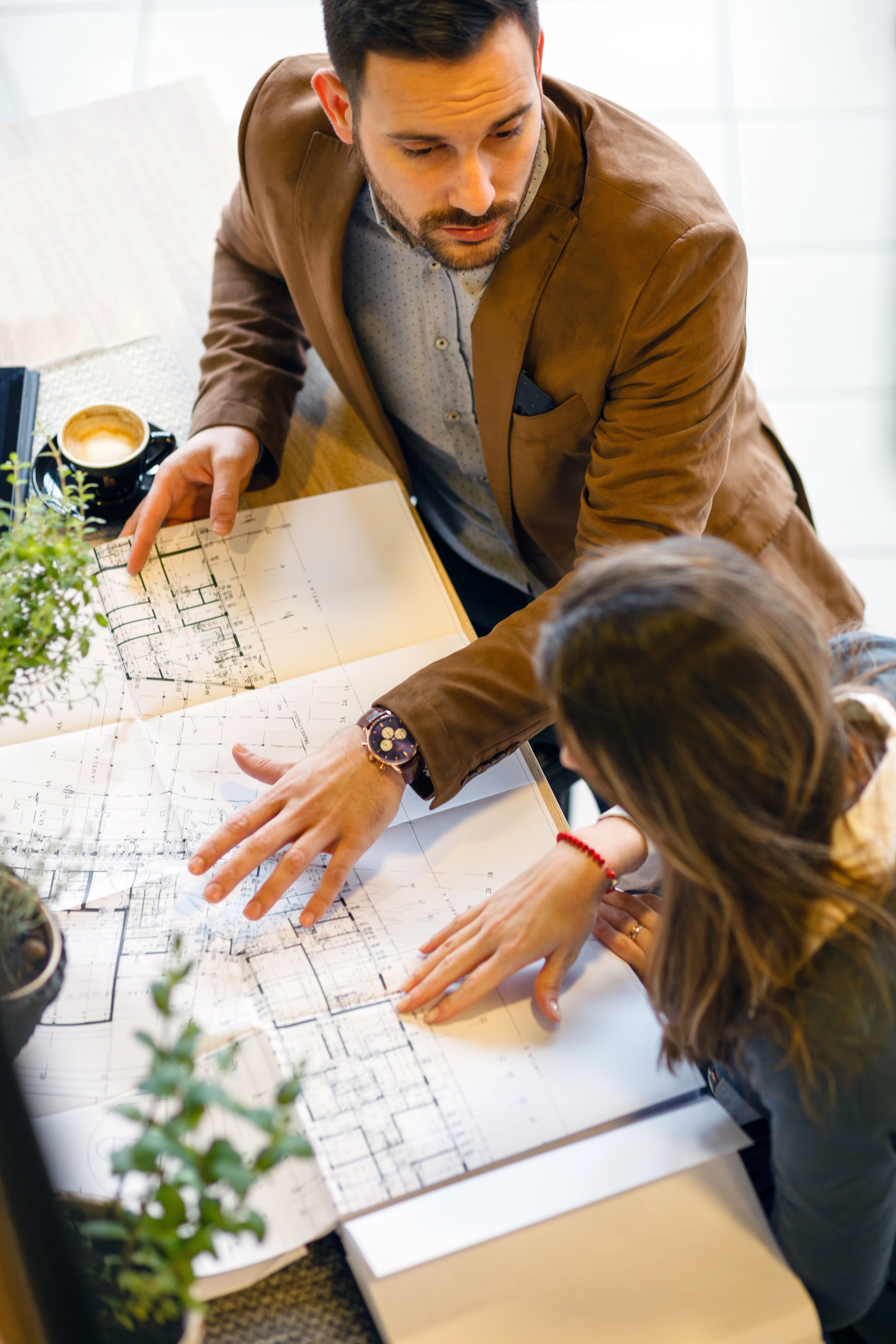 Two people, a man and a woman, examining blueprints or architectural plans on a table, with a coffee cup and plants nearby.