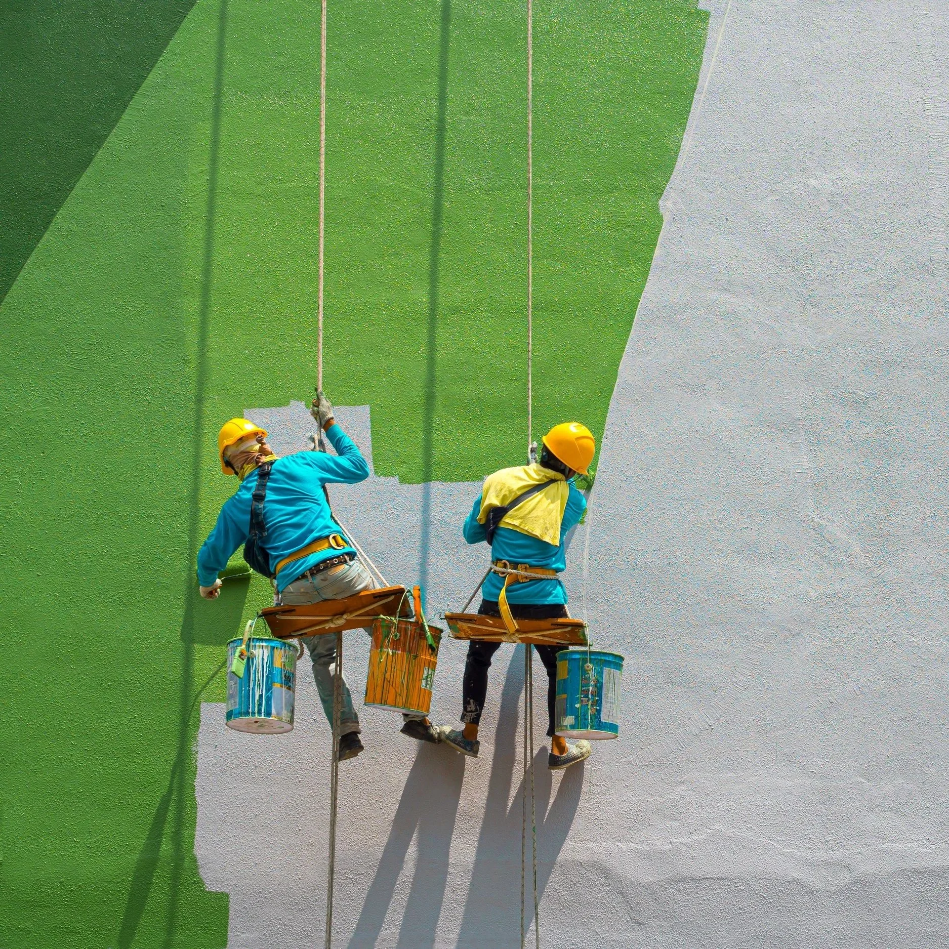 Two mural artists working on a large wall, painting a green background with gray sections.