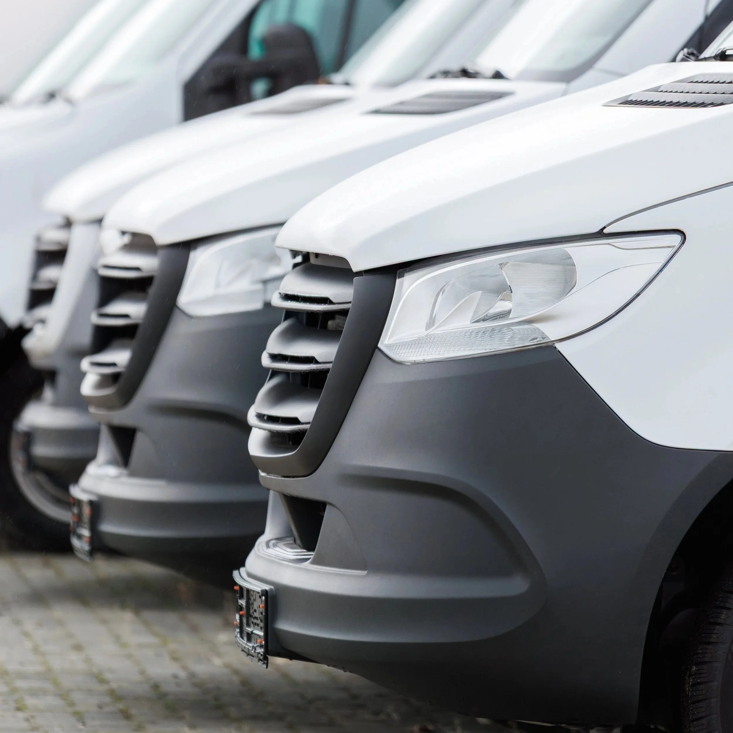 Close-up of white cargo vans parked in a row, showing front grilles and headlights.
