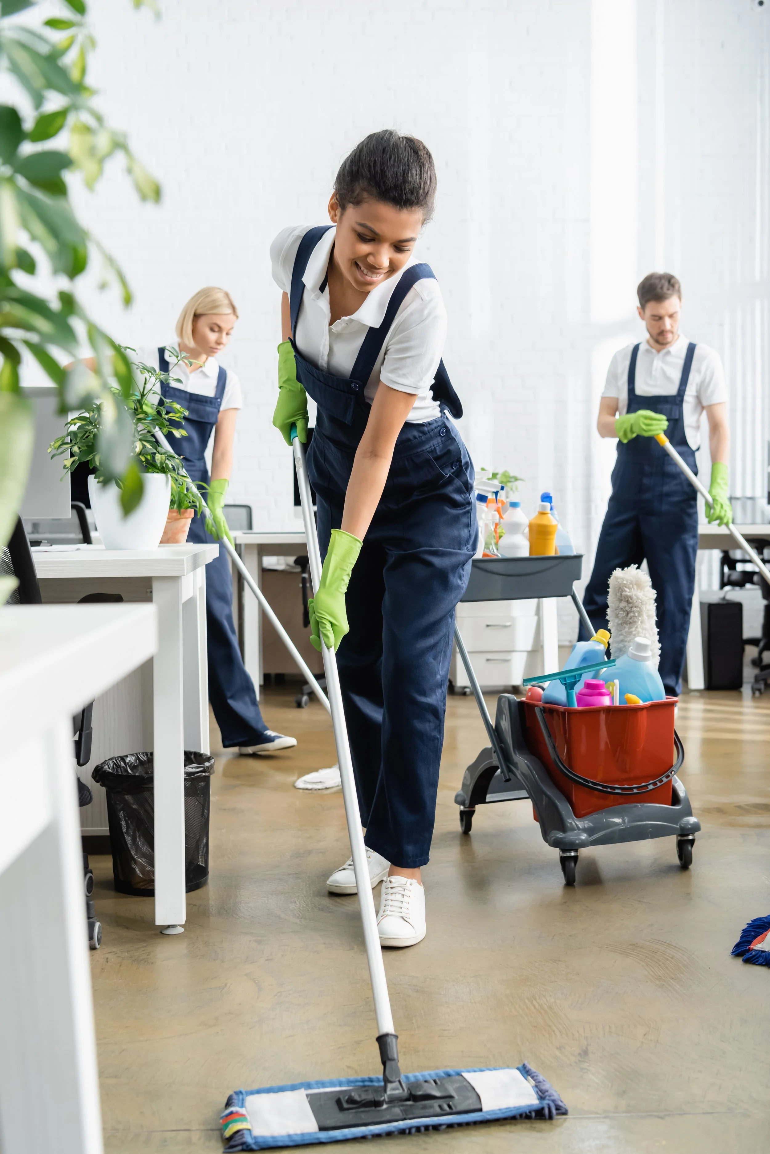 Three people in cleaning uniforms cleaning an office with mops, one woman in the foreground smiling as she mops the floor, with cleaning supplies nearby.