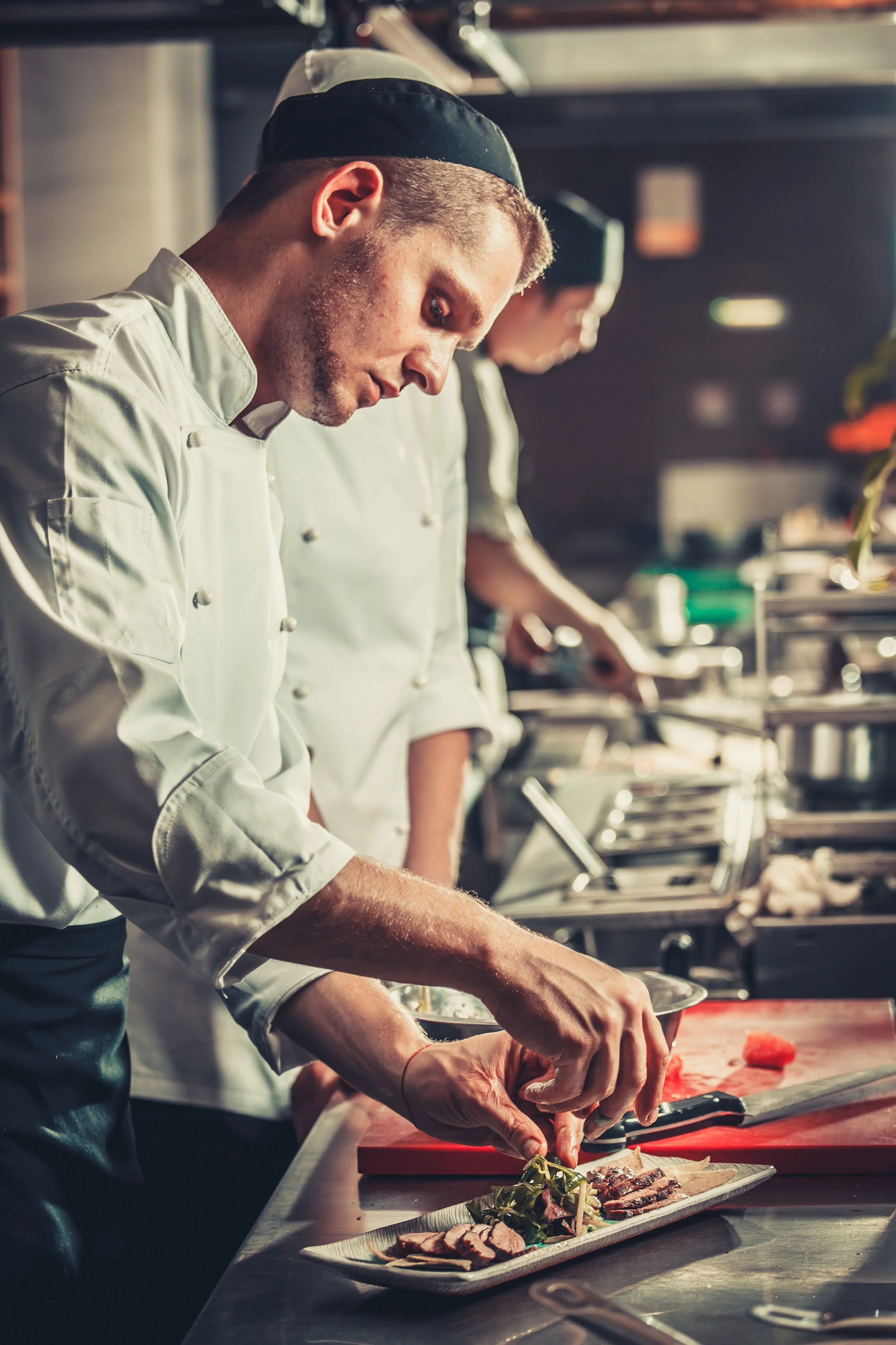 A chef garnishing a plated dish in a professional kitchen, with another chef working in the background.