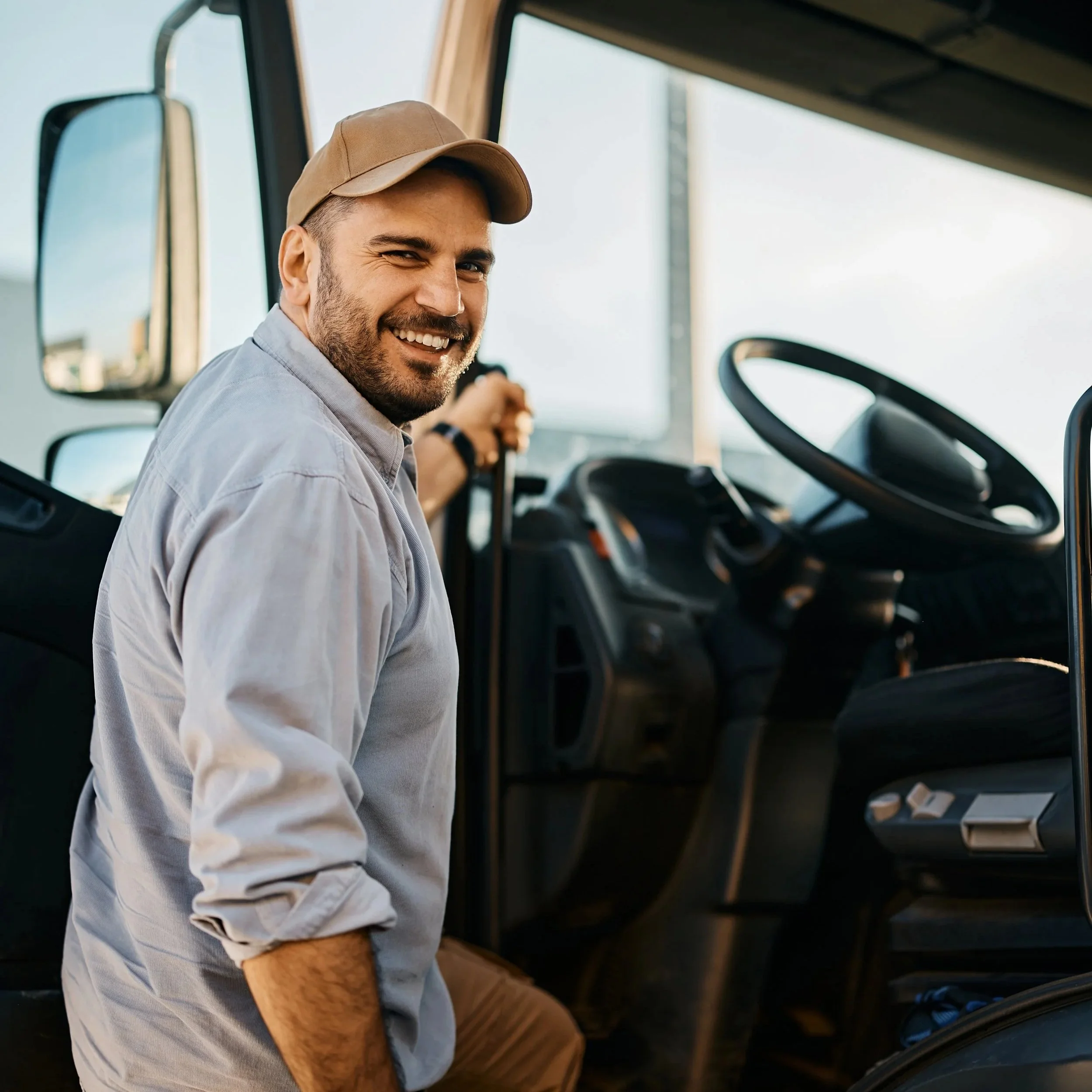 Smiling man in a beige cap and light gray shirt standing inside a vehicle, holding a steering wheel.