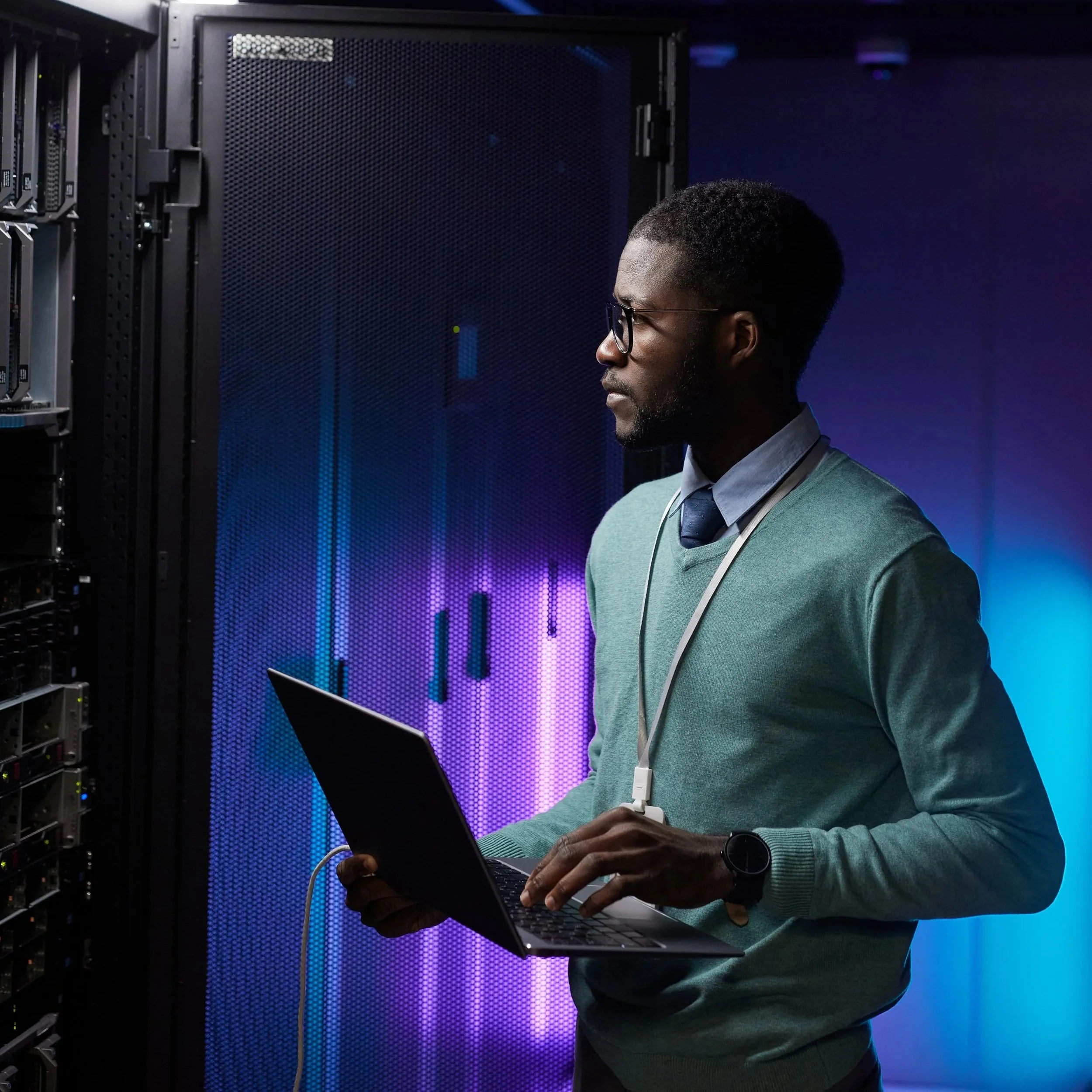 A man working in a data center, holding a laptop and standing near server racks with purple and blue lighting.