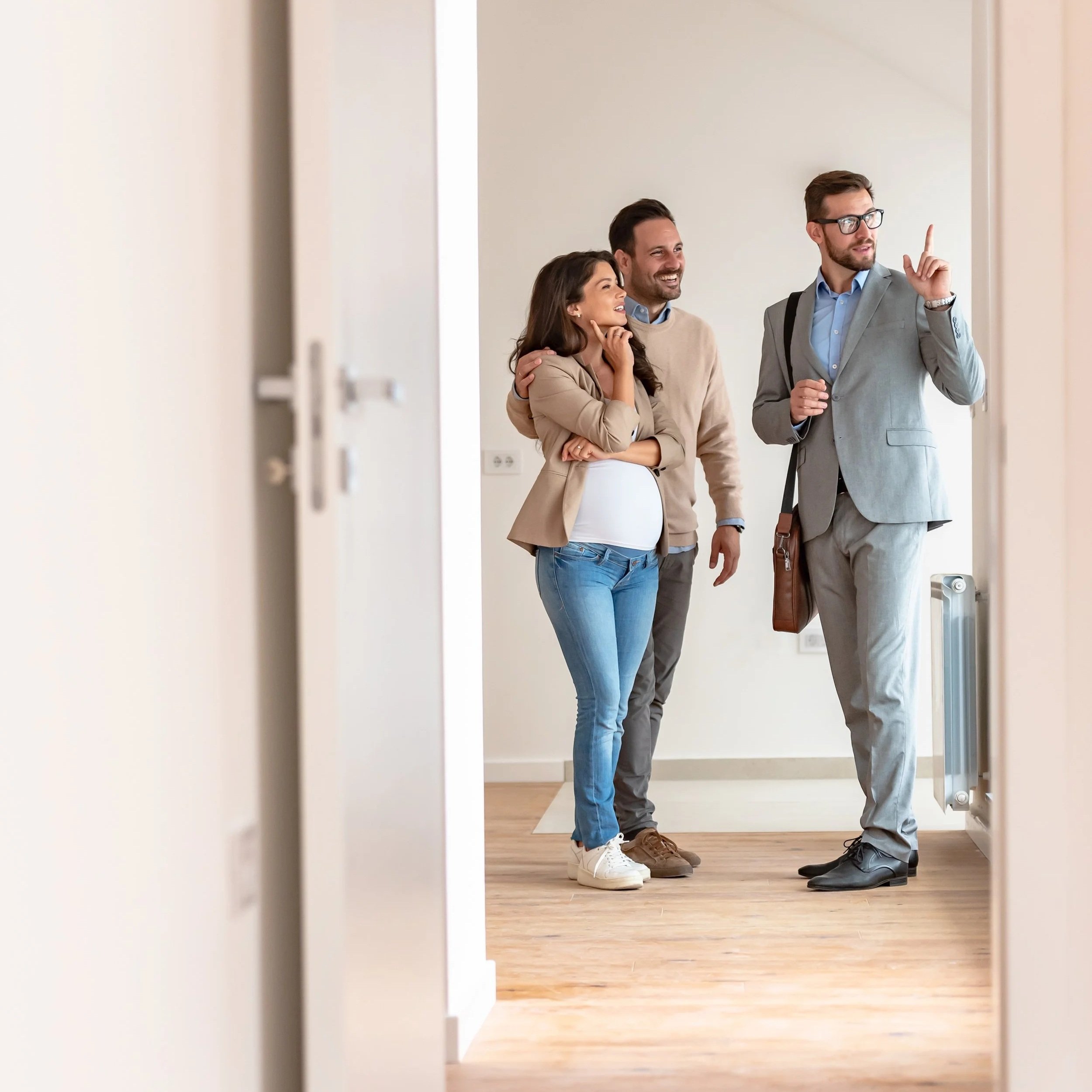 Three people, a woman and two men, are standing in a hallway of an apartment or office building. The woman appears to be pregnant. They are engaged in conversation, with one man gesturing as if explaining something.