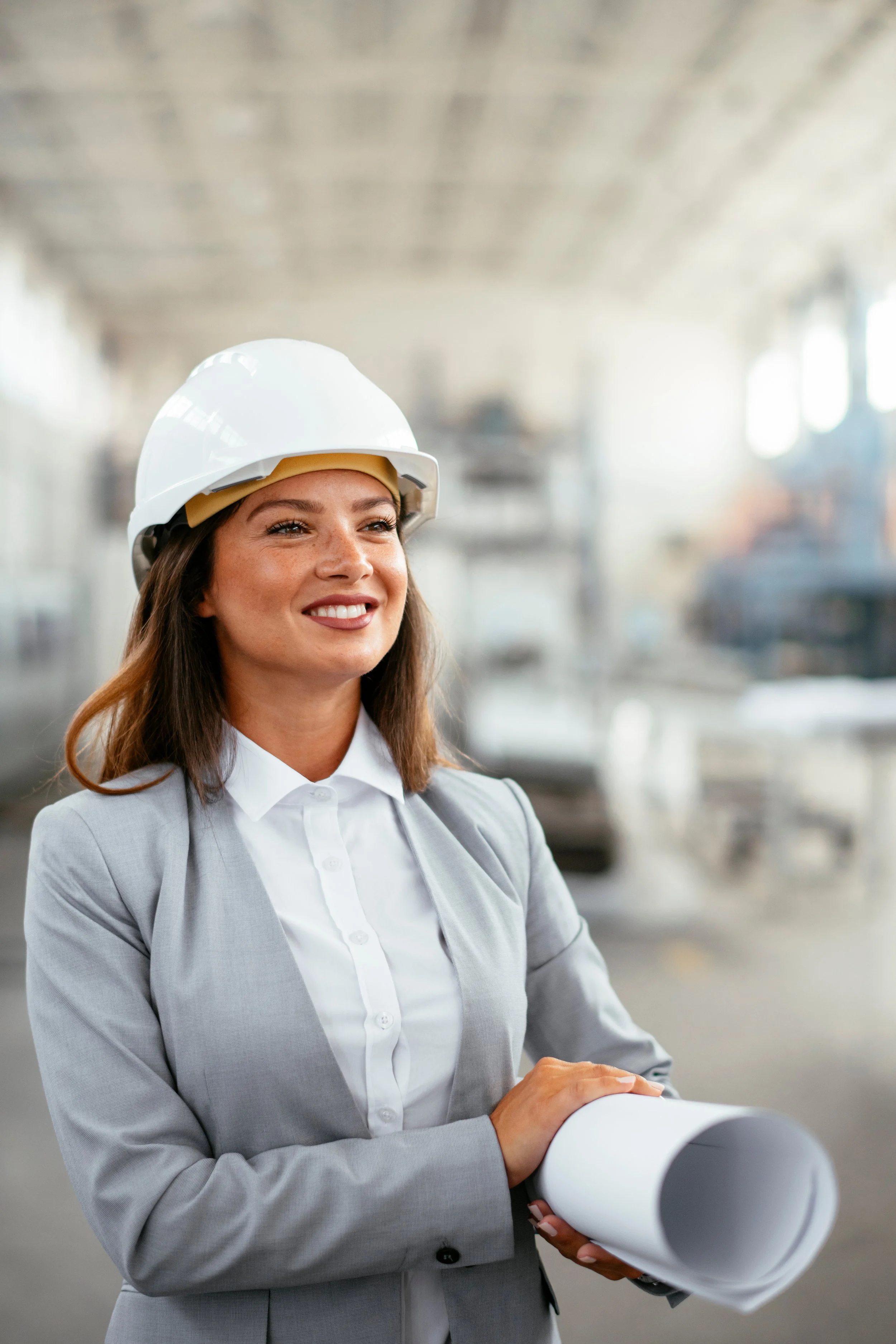 A smiling woman in a gray blazer and white shirt wearing a white safety helmet, holding rolled-up blueprints in an industrial setting.