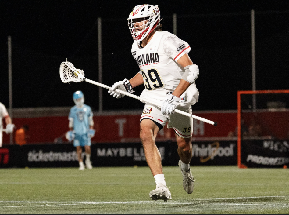 Lacrosse player in a white and red uniform with a pink helmet, holding a lacrosse stick, near the goal in a game.
