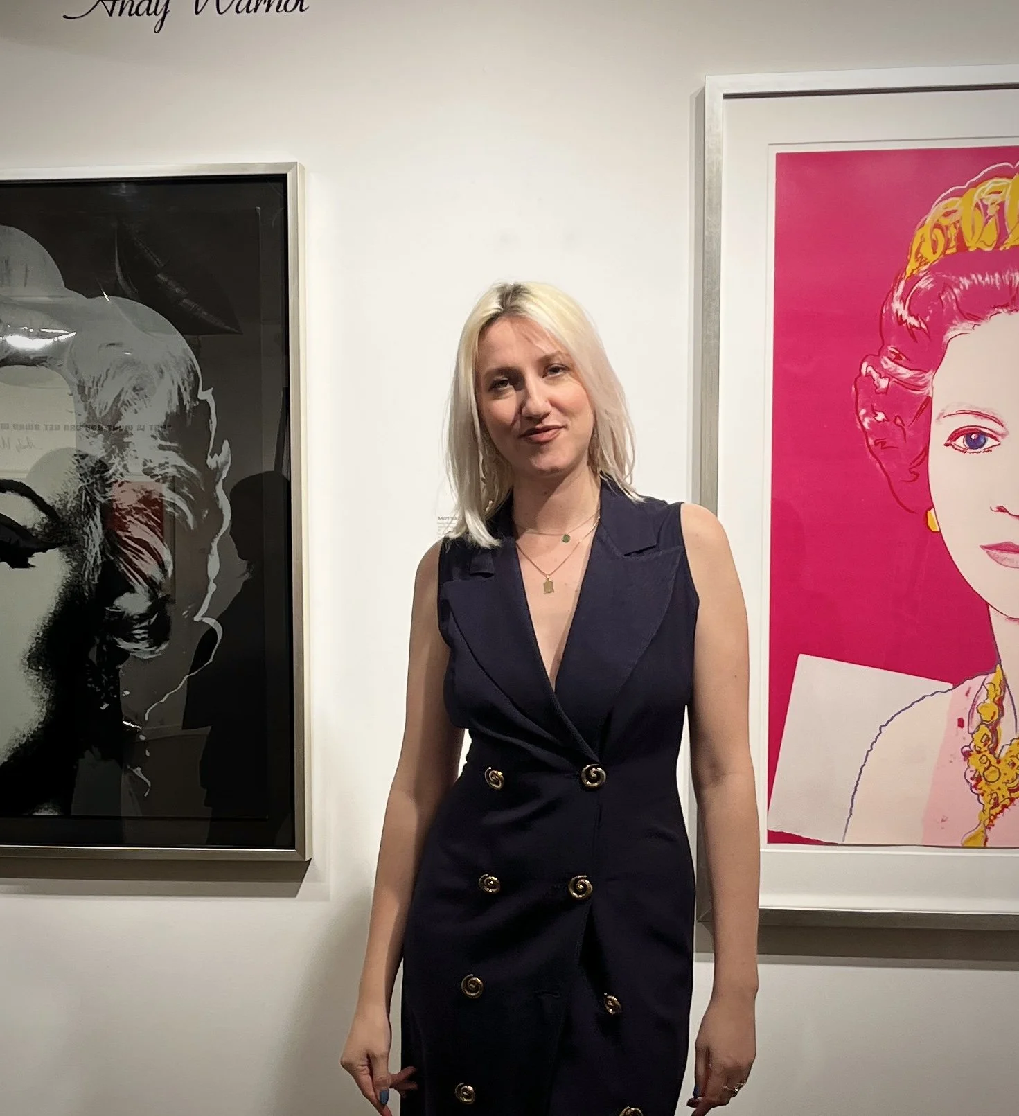 A young woman (Jelena Jankovic) with blonde hair wearing a dark navy sleeveless dress with gold spiral buttons standing in front of an artwork by Murakami on a white wall.