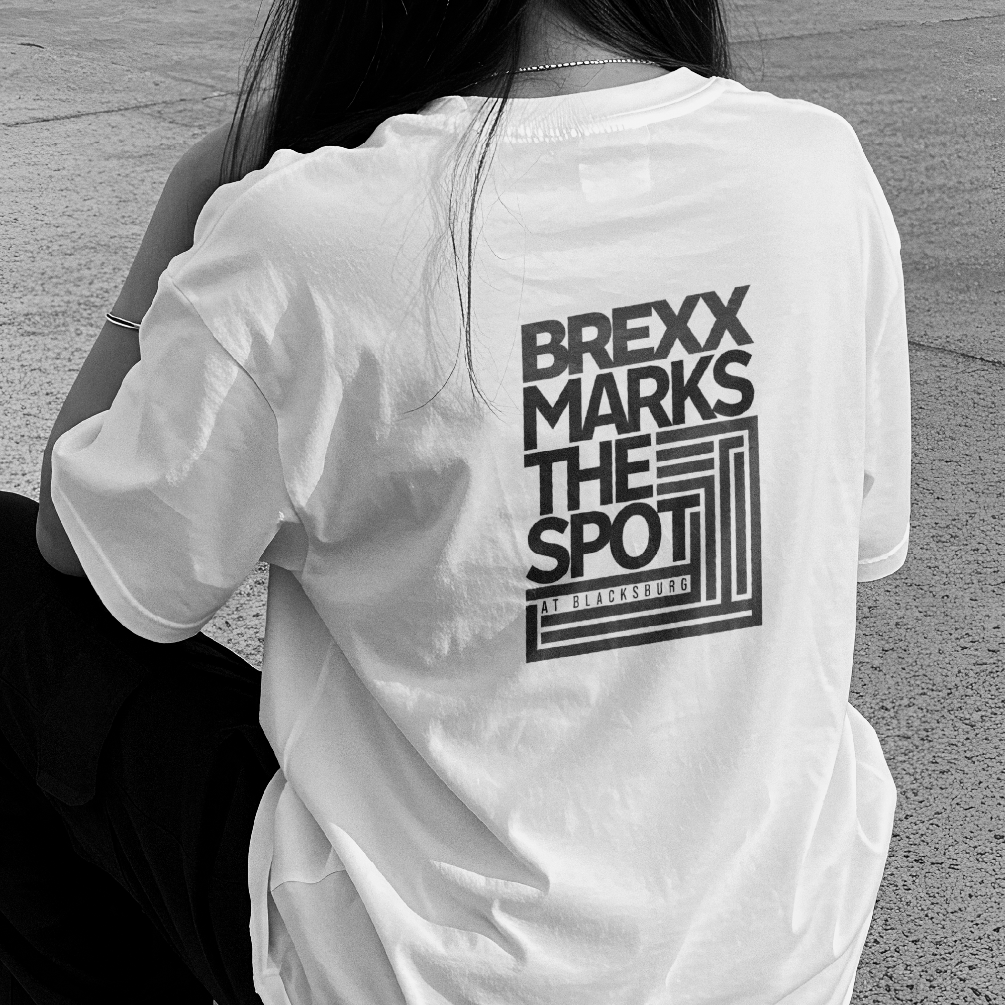 Back of person wearing a white T-shirt with black text, sitting outdoors on pavement, with long dark hair and a bracelet on their wrist.