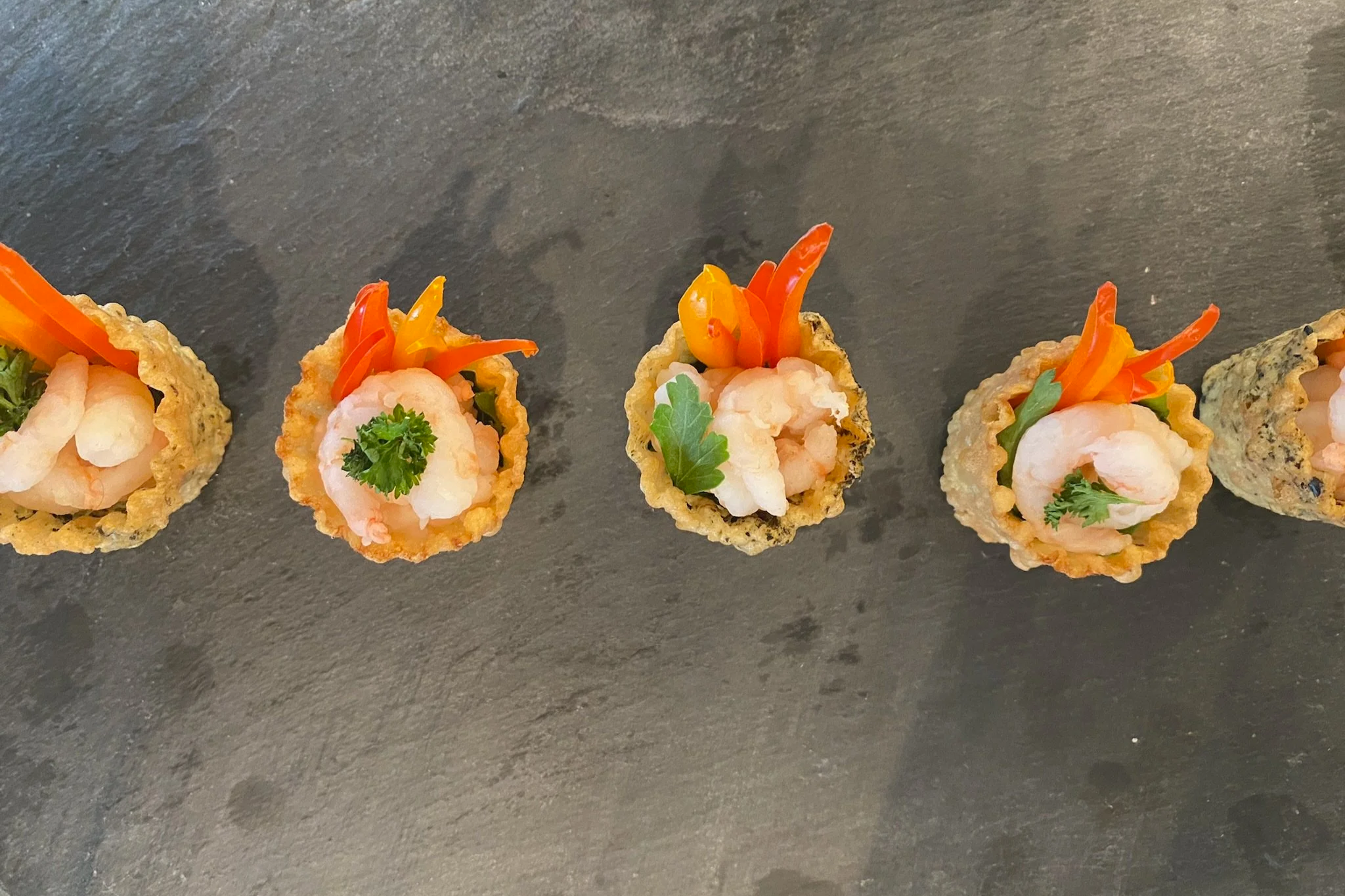 Close-up of five shrimp canapés with colorful vegetables and herbs on a dark surface.