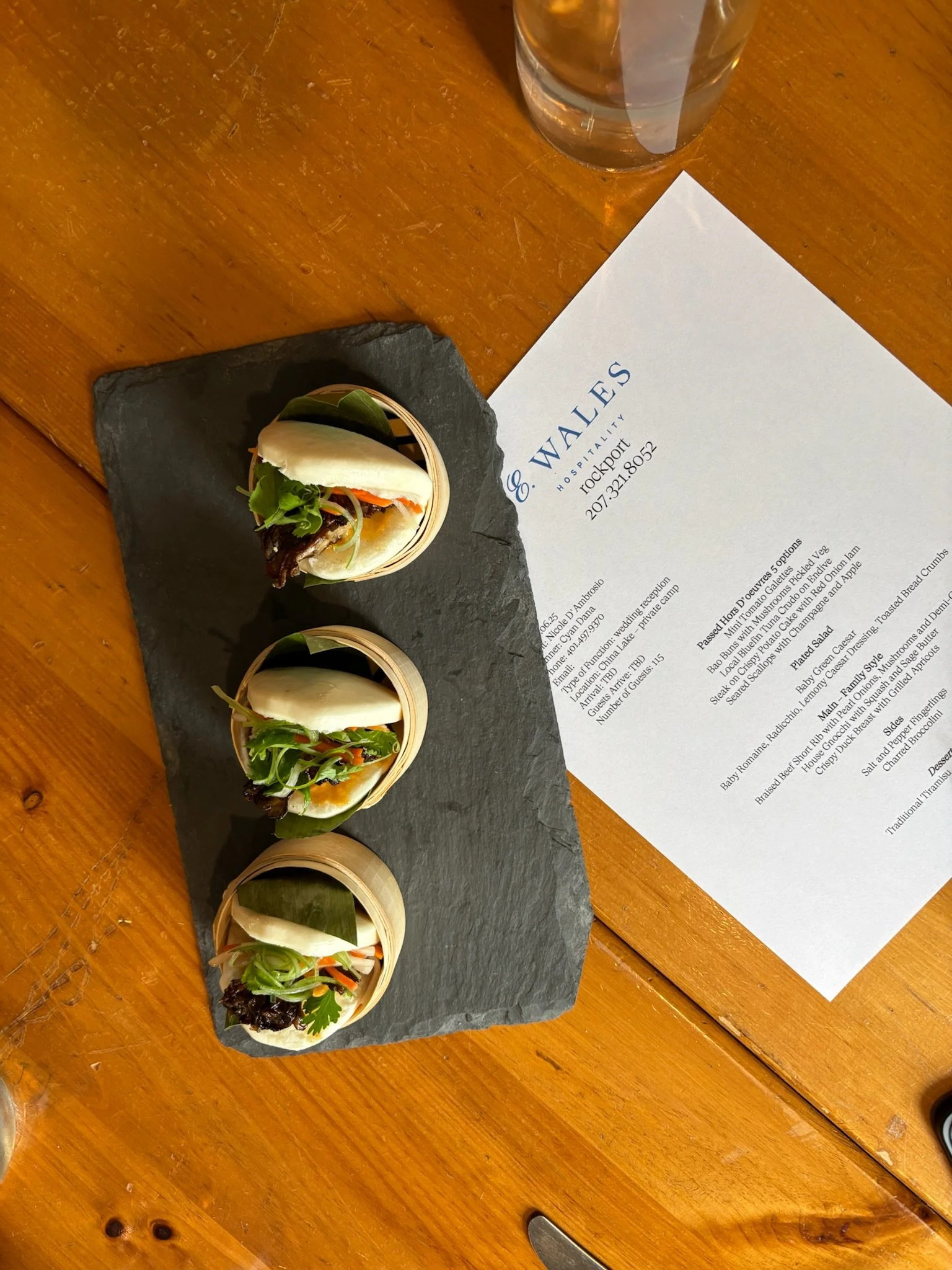 Three small bamboo steamers with sandwiches on a black slate serving tray, a glass of water, and a menu on a wooden table.