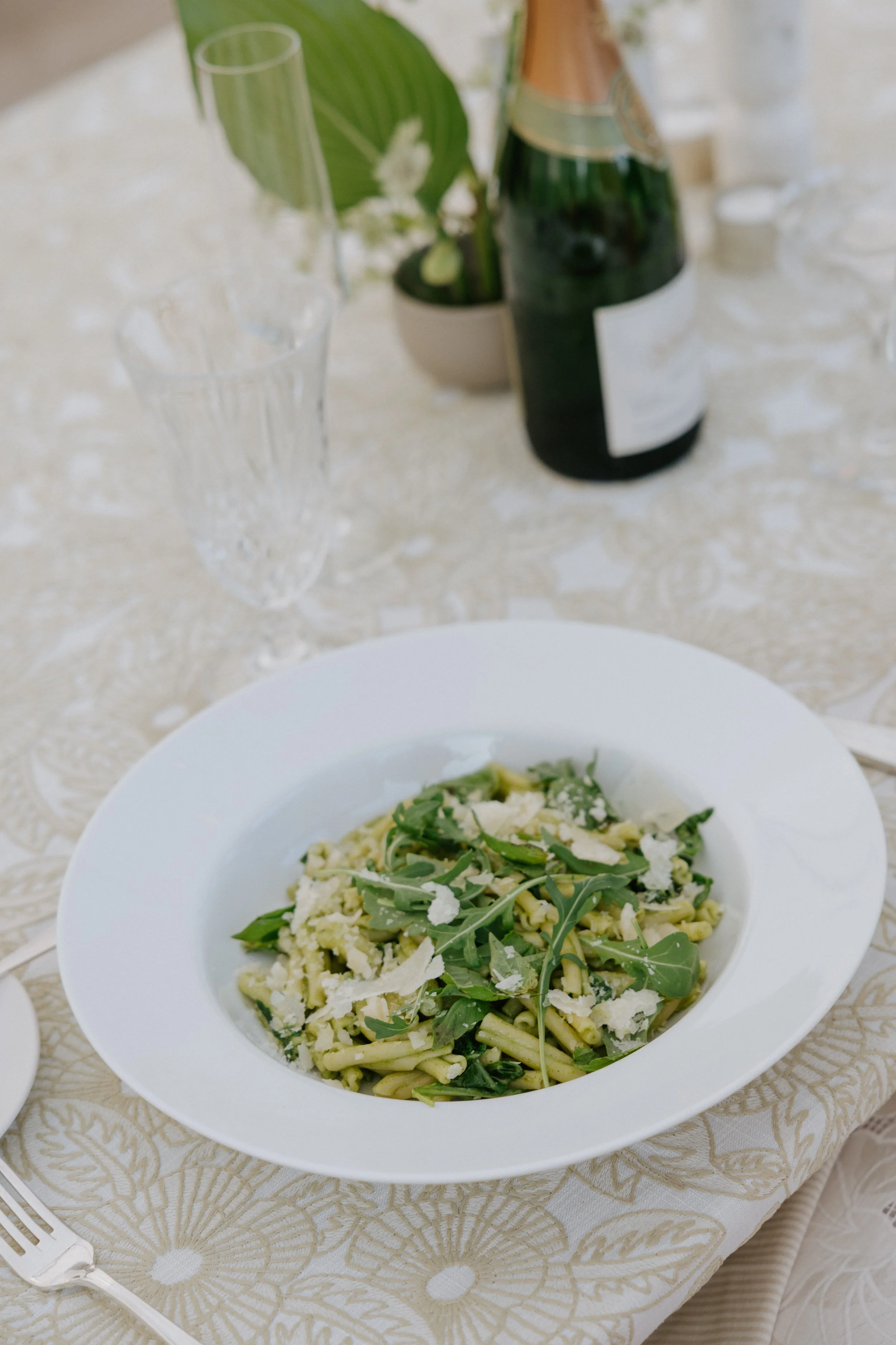 A plate of pasta with arugula and grated cheese on a table set for a meal. There is a glass of champagne and a bottle of champagne in the background, along with a small potted plant.