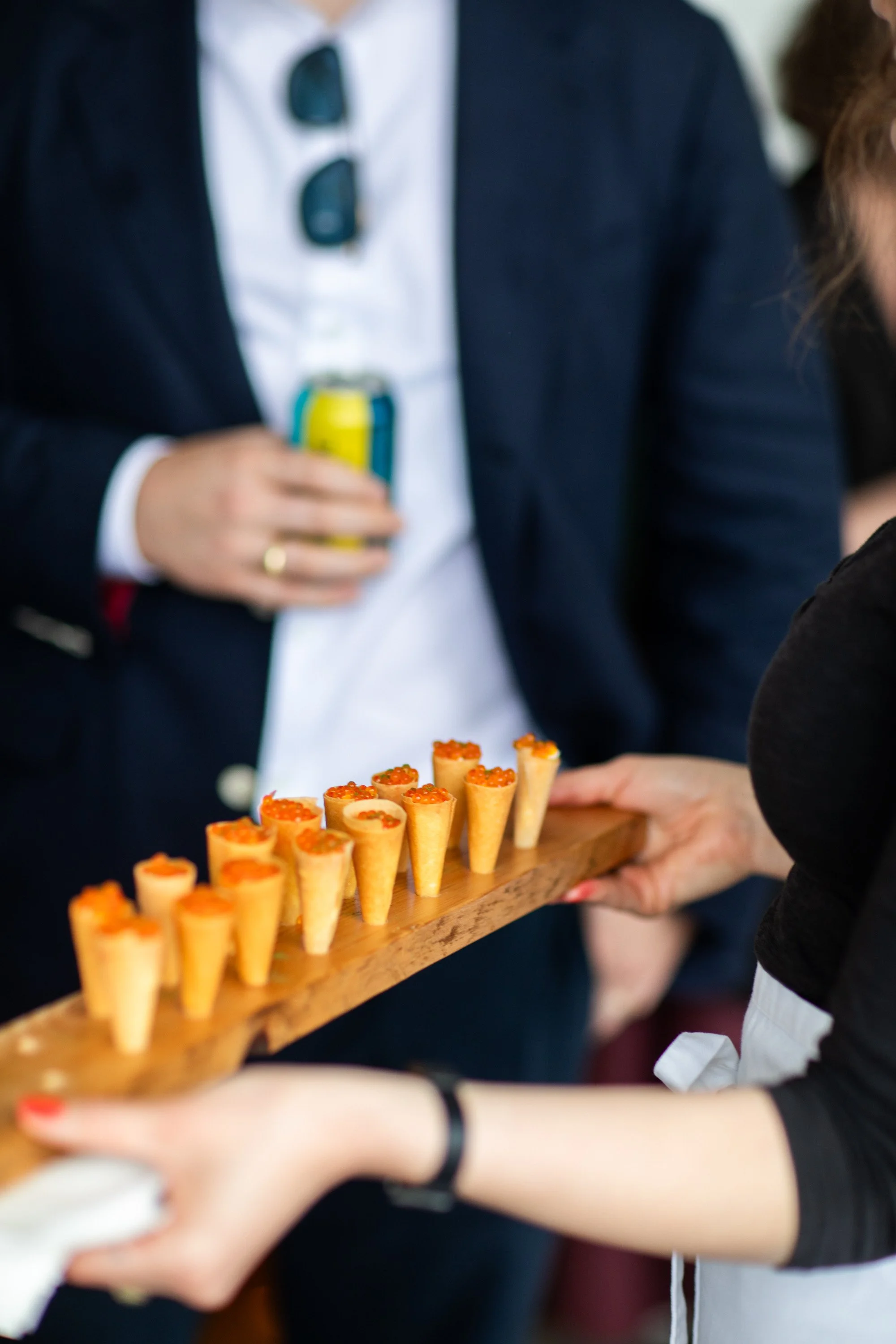 A person holding a wooden tray with small cone-shaped appetizers topped with orange fish roe. In the background, a person wearing a dark jacket and sunglasses hanging from the collar, holding a can.