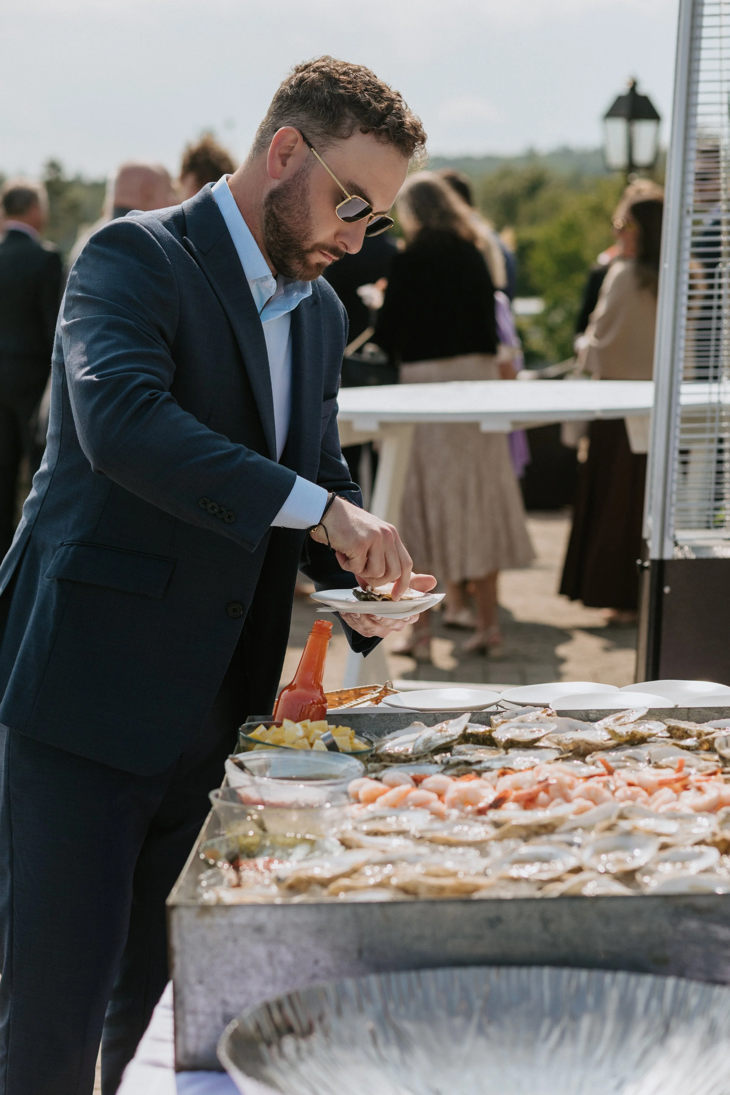 A man in a suit with sunglasses serving himself seafood at an outdoor buffet table during an event.