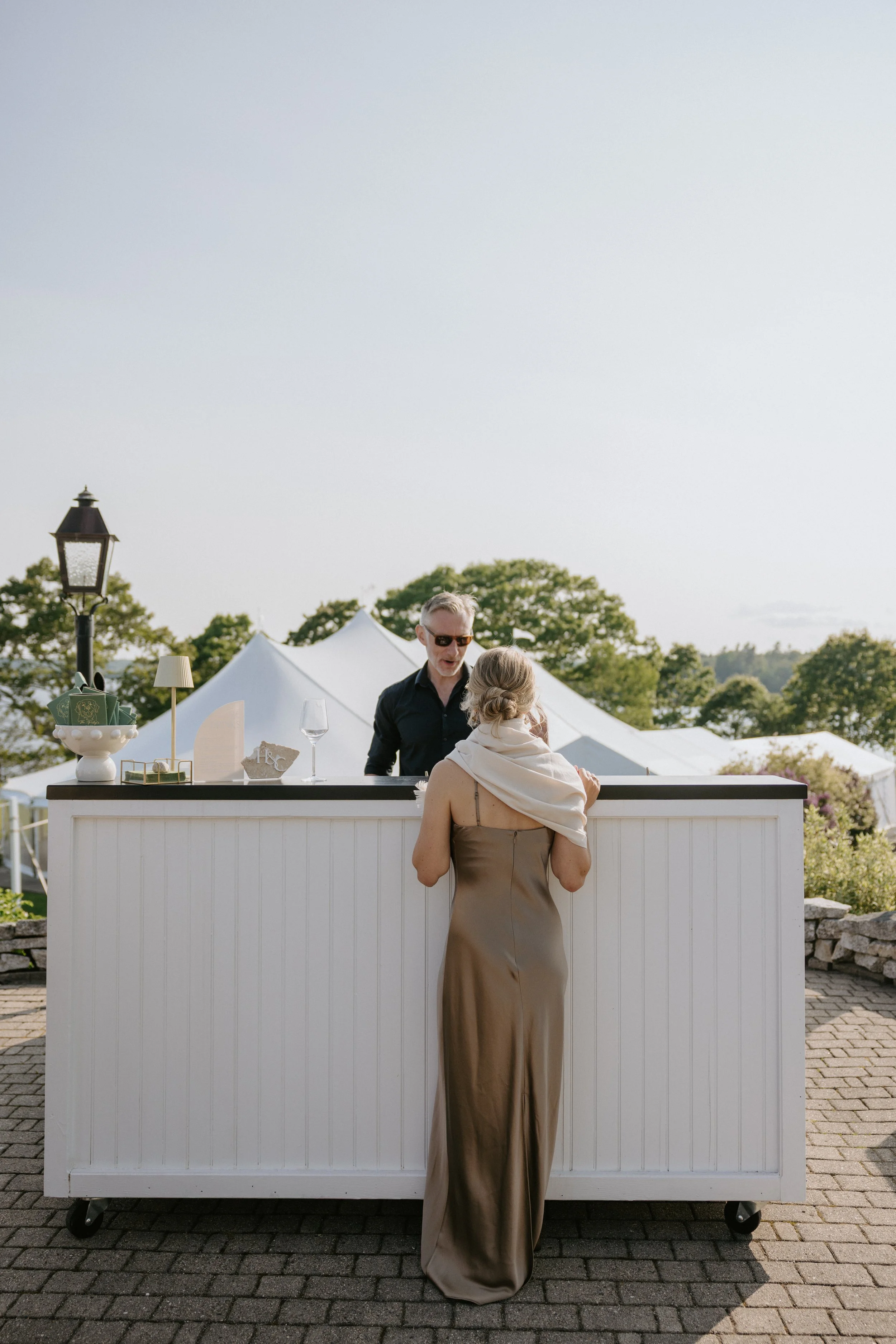 A woman in a brown silk dress with a white scarf around her shoulders is standing at an outdoor bar, speaking with a man who is behind the bar. The scene is set outdoors with trees, tents, and a stone wall in the background, during the daytime.