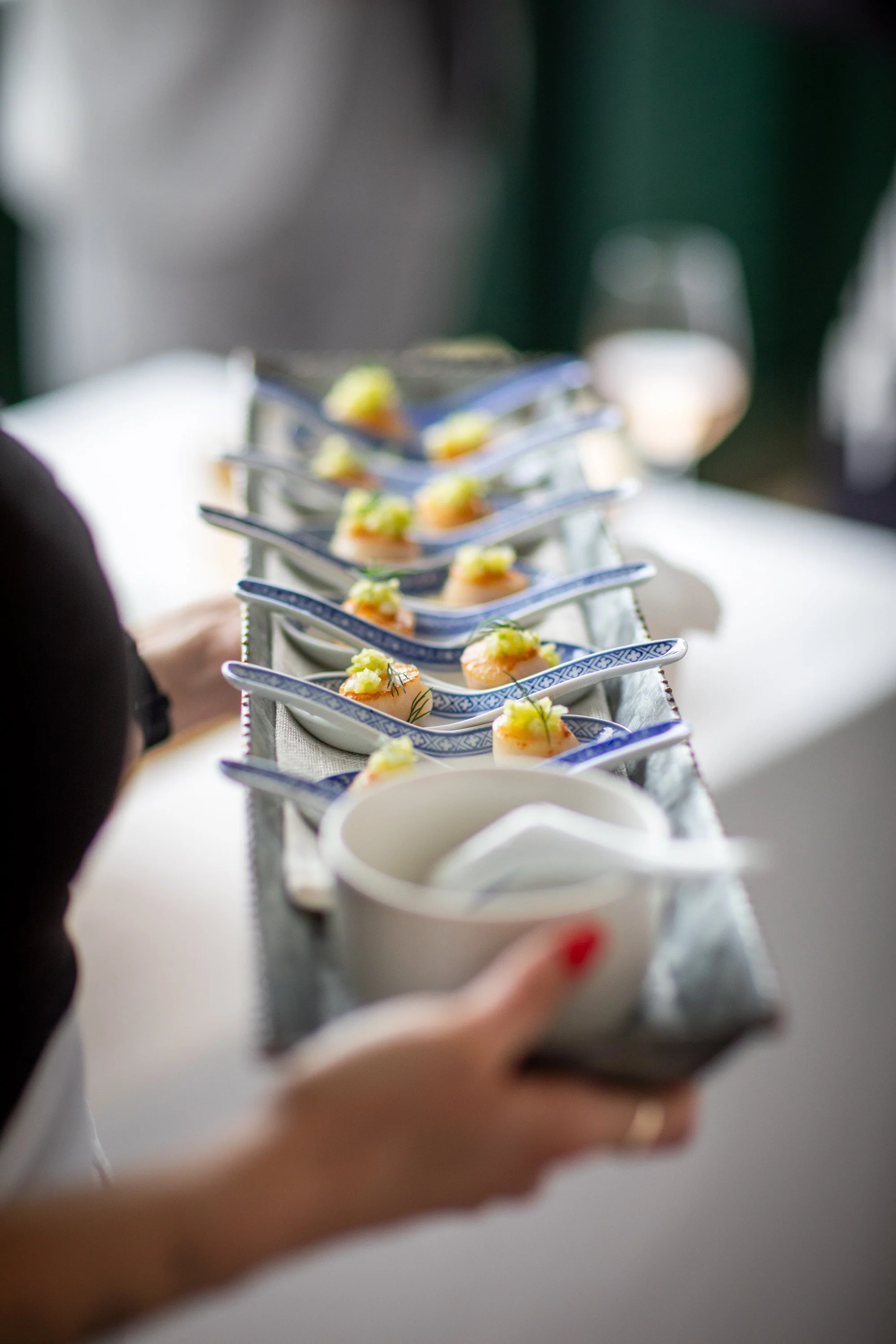 A server holding a tray of small appetizers, each on a ceramic spoon, garnished with herbs and lemon, with a glass of white wine in the background.