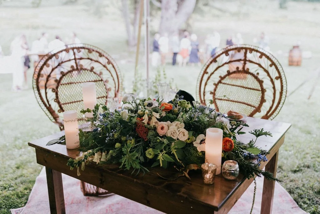 Decorated table with candles and a floral arrangement outdoors, with chairs and people in the background.