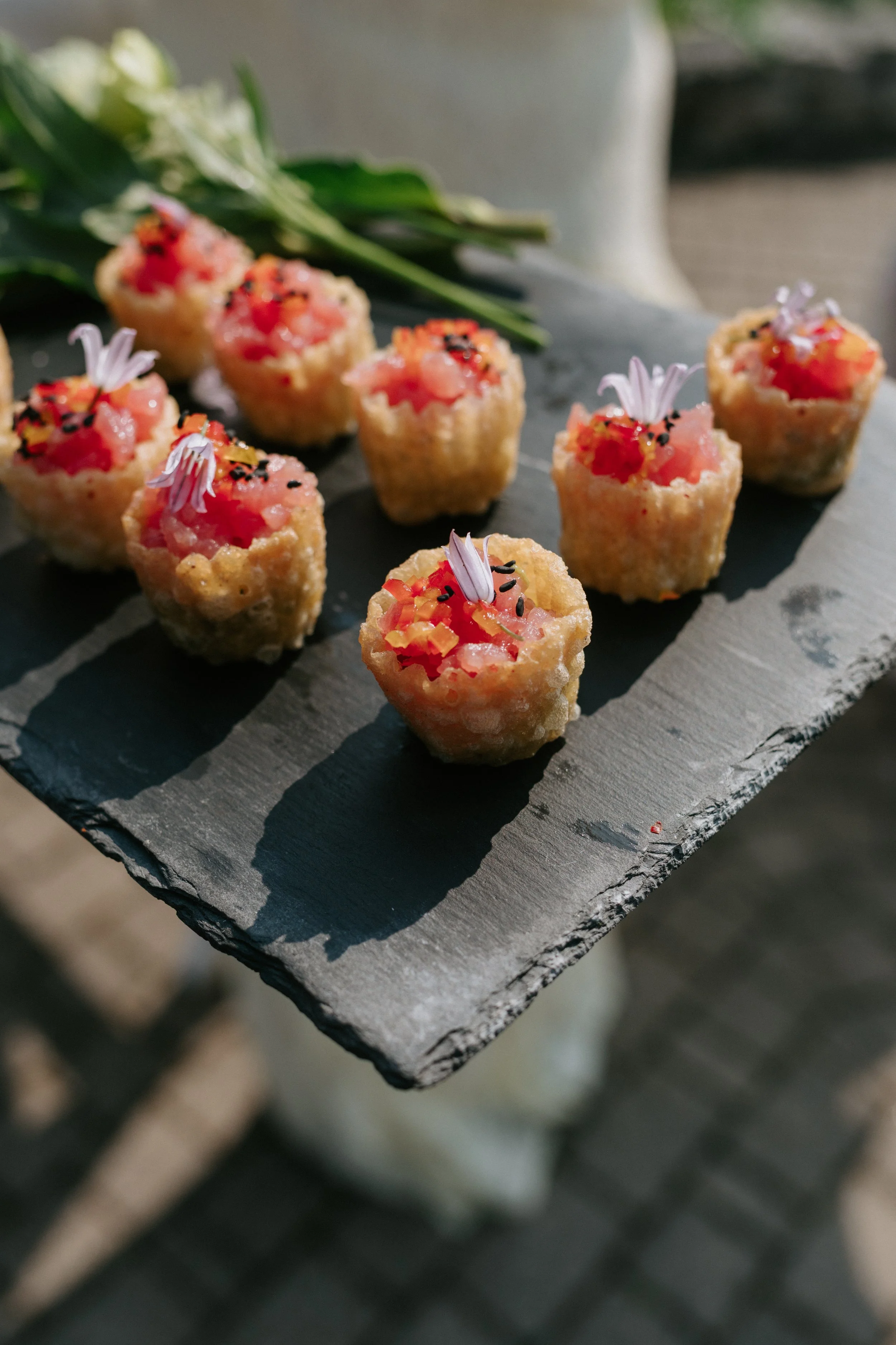 Eight small appetizers with pink topping, garnished with tiny edible flower petals and black sesame seeds, arranged on a black slate serving board.
