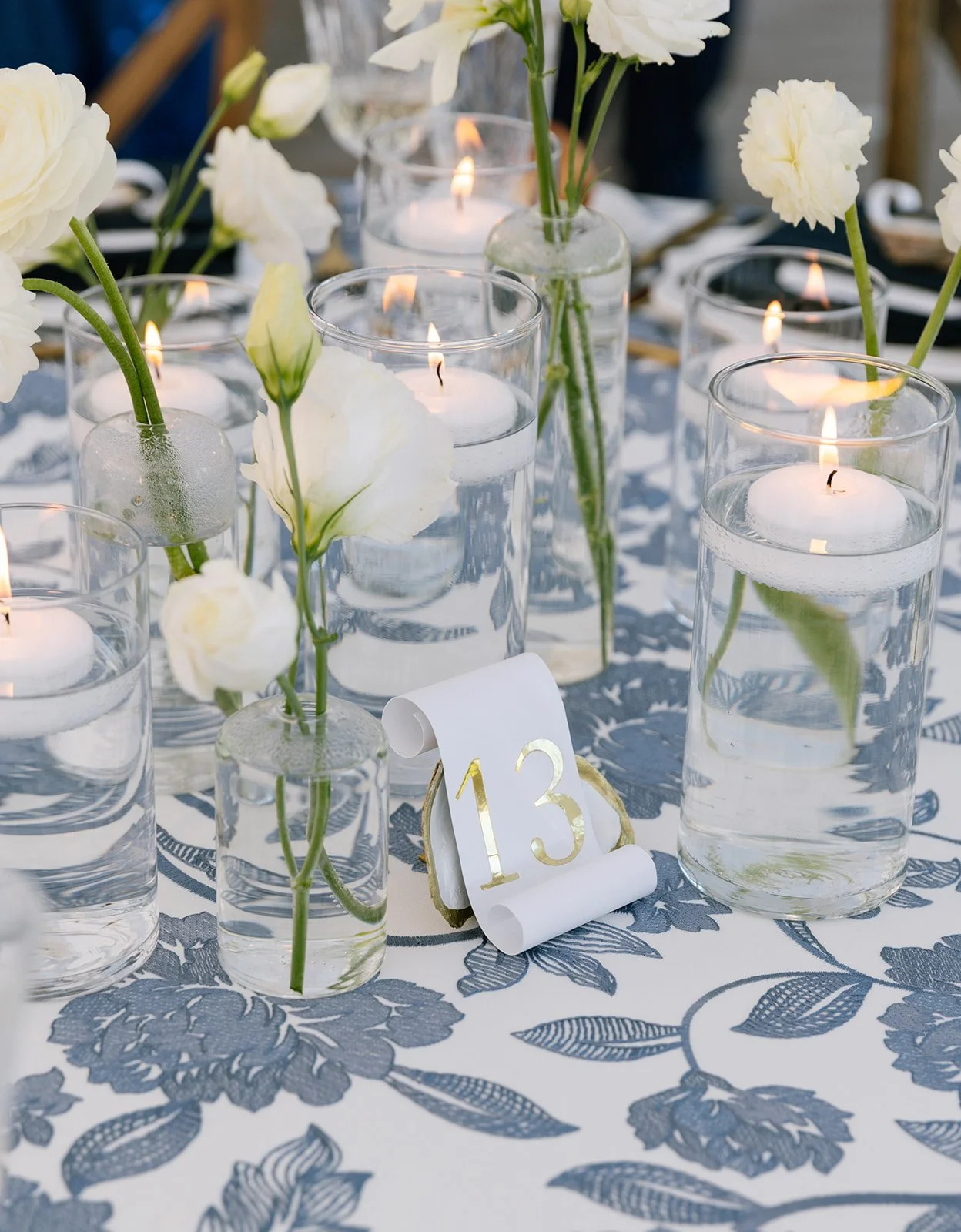Decorated table with candles and a floral arrangement outdoors, with chairs and people in the background.
