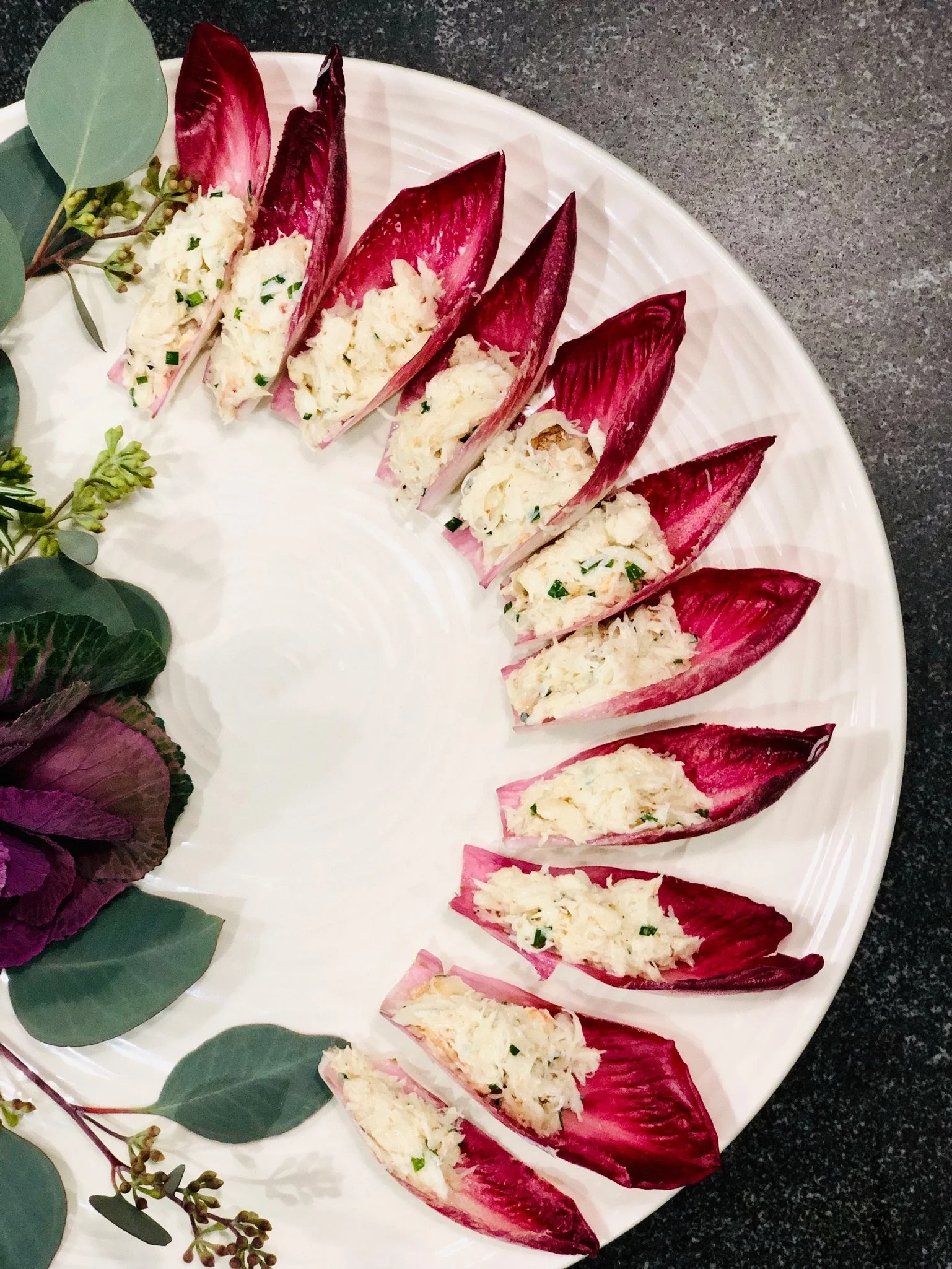 Radicchio leaf boats filled with a crab or seafood salad, arranged in a circular pattern on a white plate with green foliage decoration on the side.