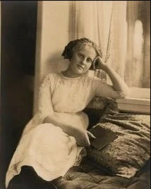 A woman seated on a bench inside, leaning her head on her hand, with a stack of books beside her and a window with curtains in the background.