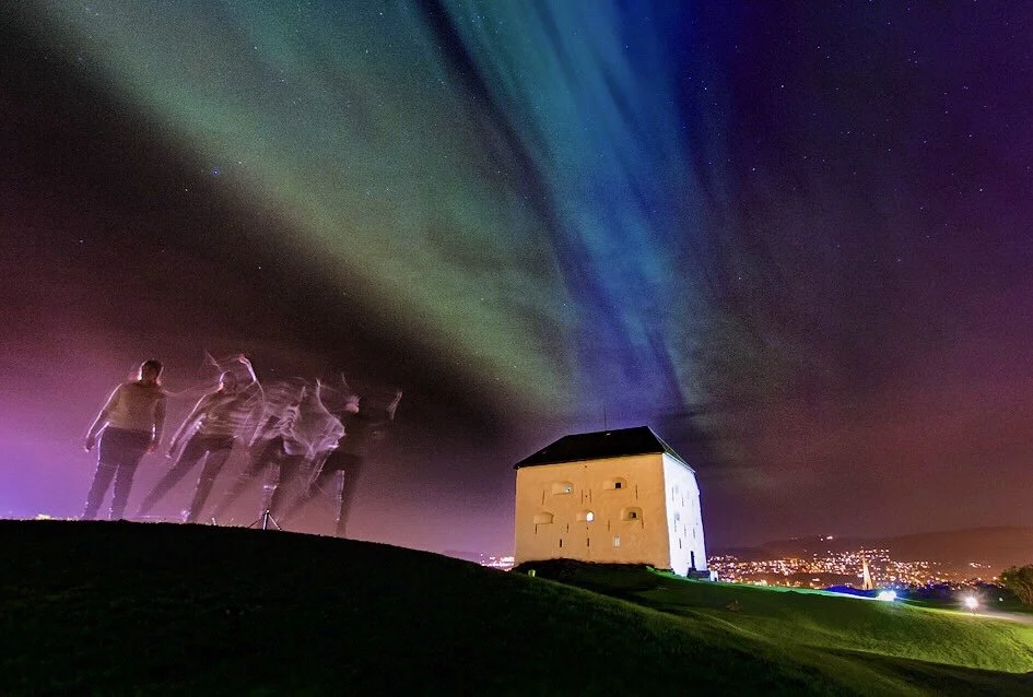 The image shows a fort from a famous Norwegian city, with northern lights above it in green and blue colors. The sky is dark purple and to the left of the fort in the foreground is a person and their ghostly dancing.