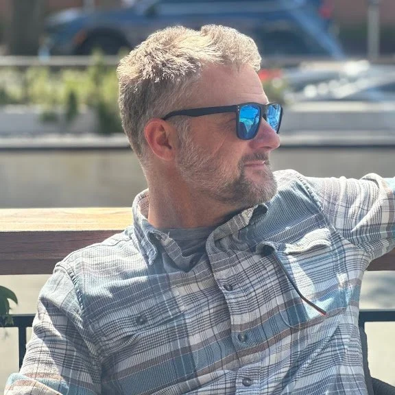 A man with short, light-colored hair and a beard, looking relaxed and confident sitting outdoors on a sunny day.