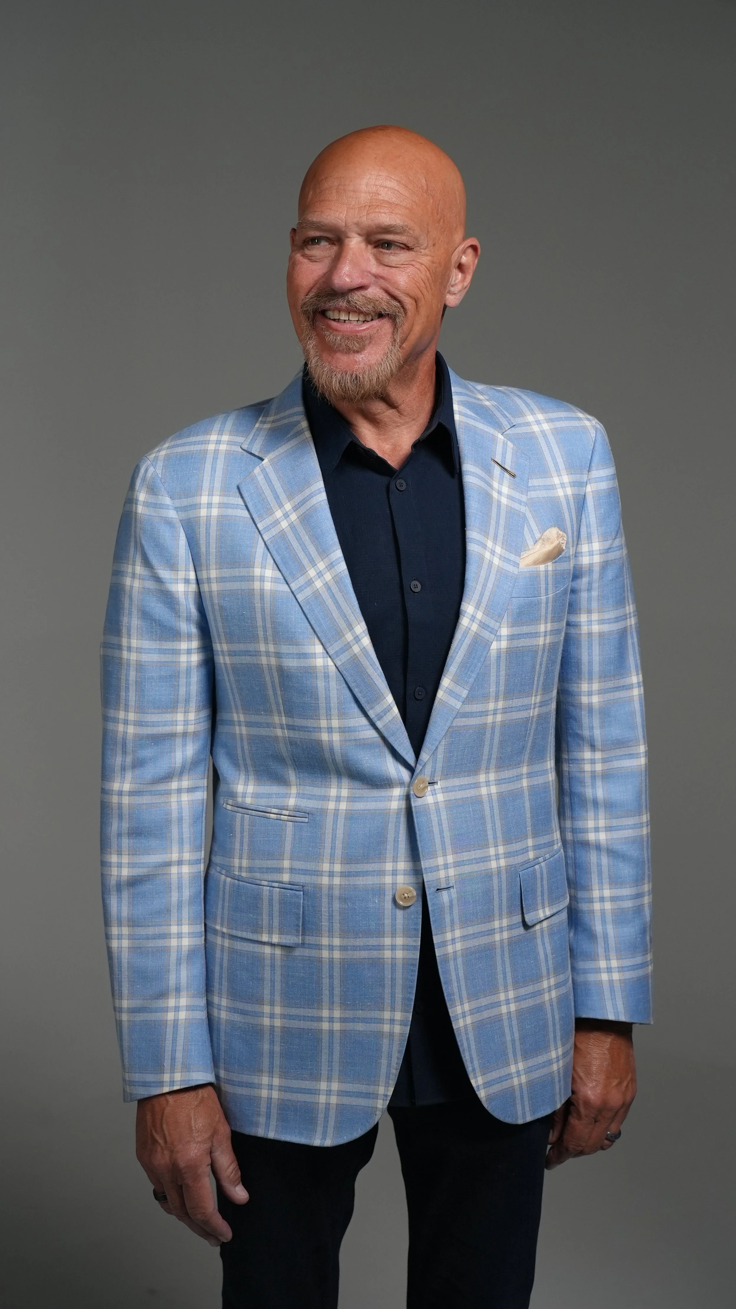 A smiling older man with a bald head and goatee, wearing a light blue plaid blazer and a dark shirt, standing against a gray background.