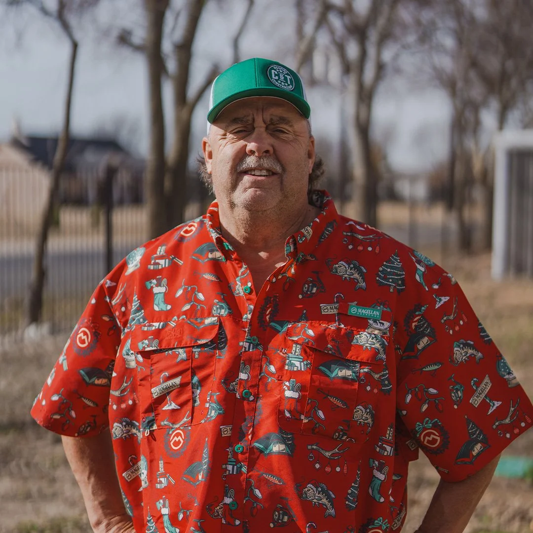 A smiling man with a mustache wearing a red and white checkered shirt, a name tag, and a truck hat, holding a small black box labeled 'Library' in front of white shutters.