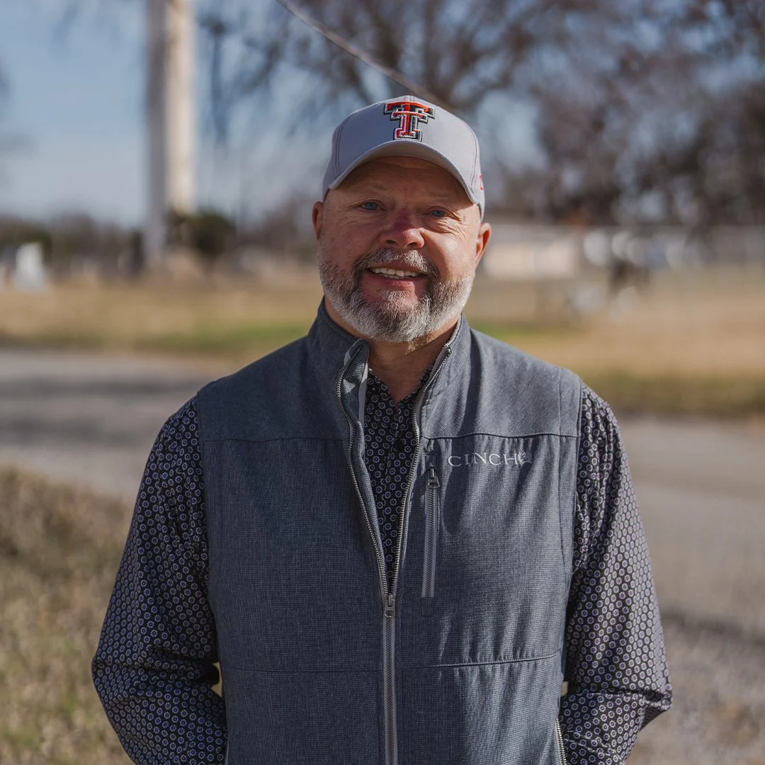 A man smiling wearing a large straw hat and sunglasses with reflective lenses, sitting outdoors at a gathering.